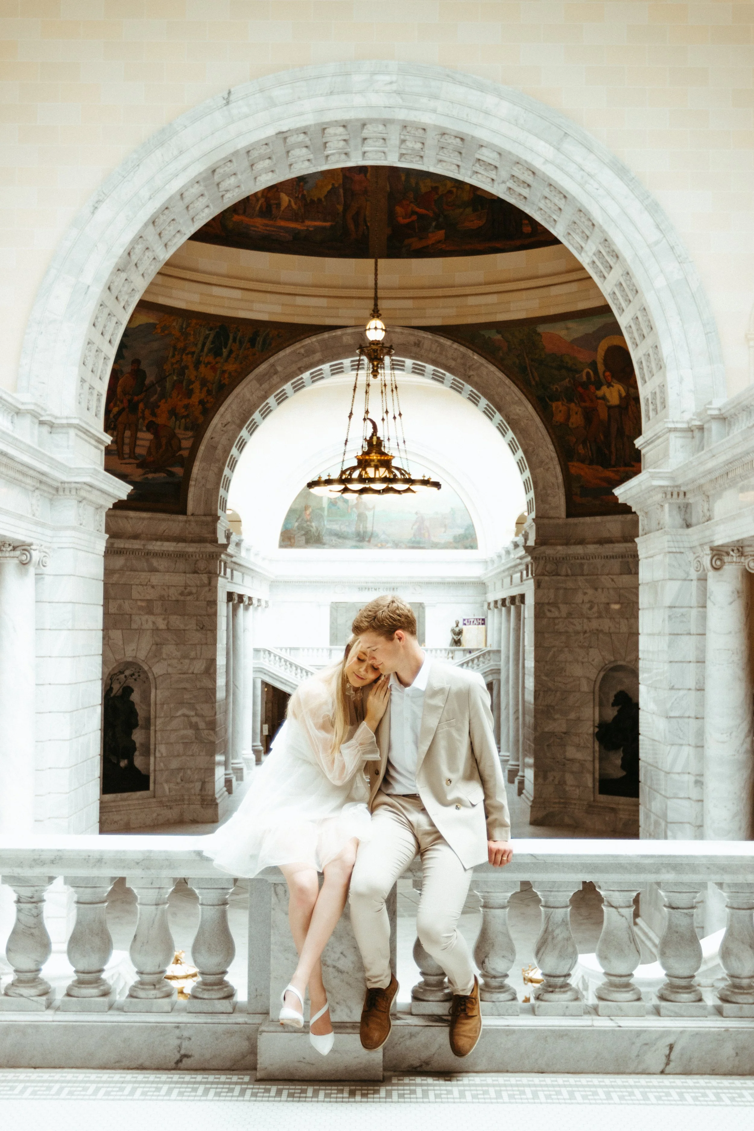 A couple dressed in wedding attire sitting on a marble railing inside a grand, historic building with arched ceilings and ornate paintings.