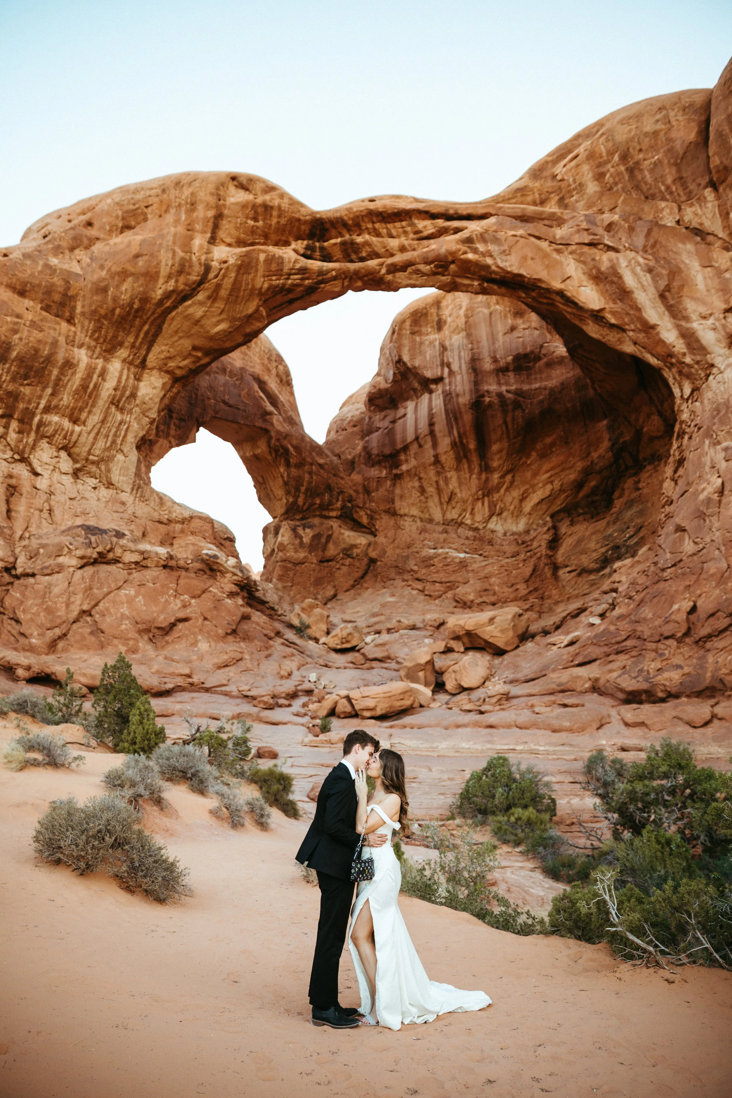 A couple dressed in wedding attire standing close together and kissing in a desert landscape with large red rock formations and two arches in the background. Arches National Park wedding photographer, Moab Utah elopement photographer. Double Arch elo