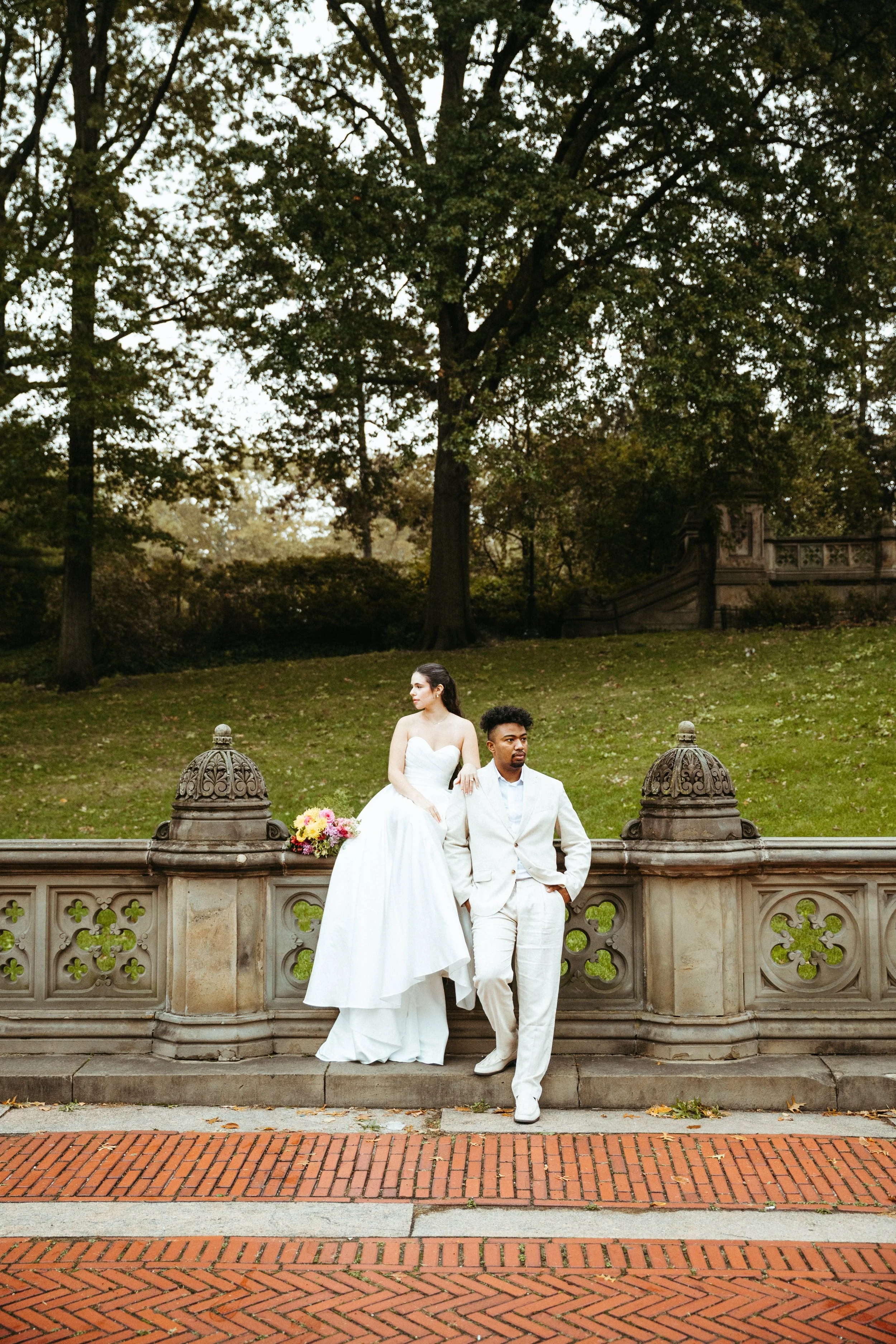 A bride in a white wedding dress and a groom in a light suit stand on a decorative stone railing in a park with large trees in the background. The bride is seated, holding a bouquet of flowers, while the groom stands with one hand in his pocket.