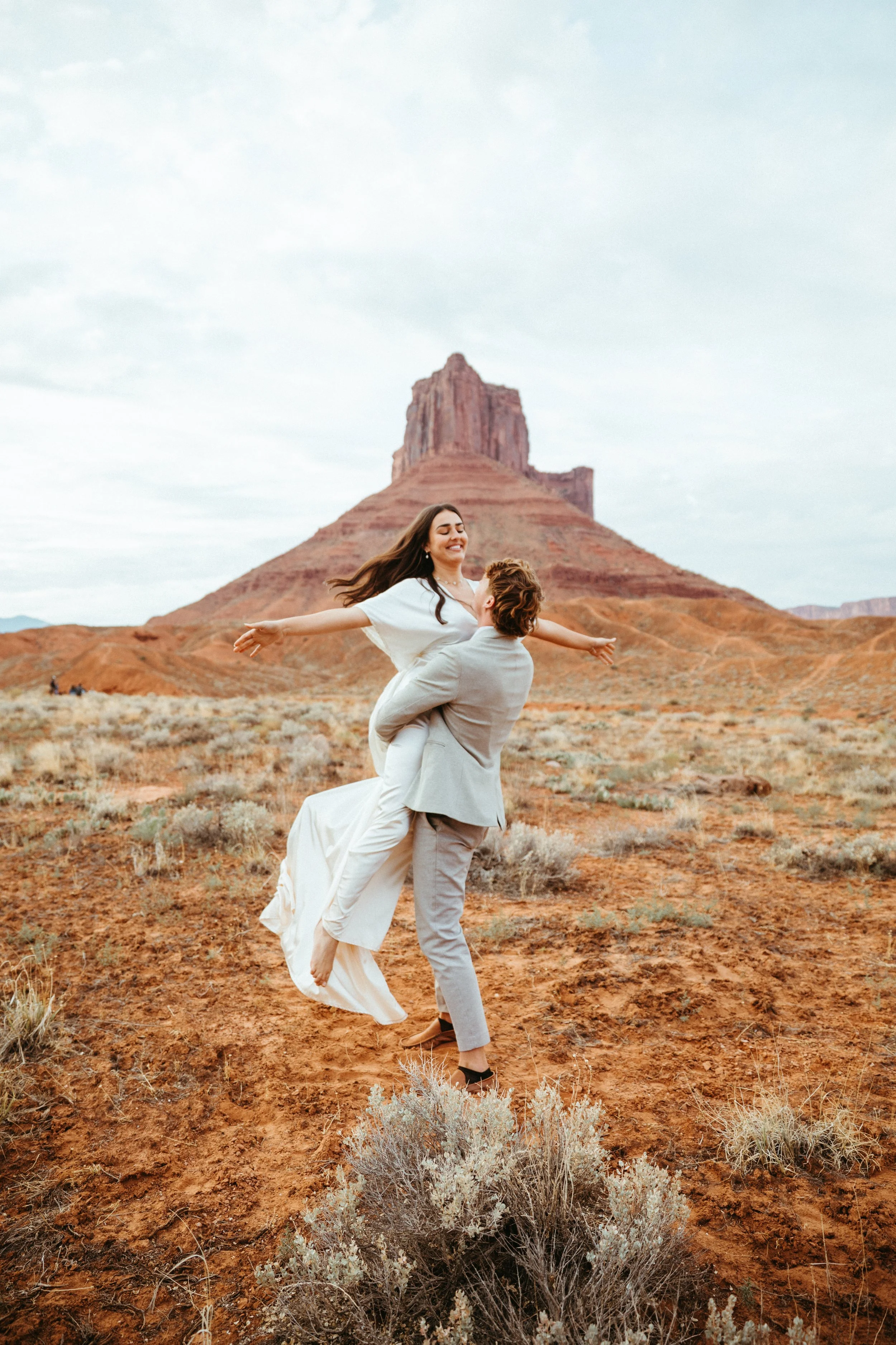 A couple in wedding attire, the man lifting the woman in a desert landscape with a large rock formation in the background.
