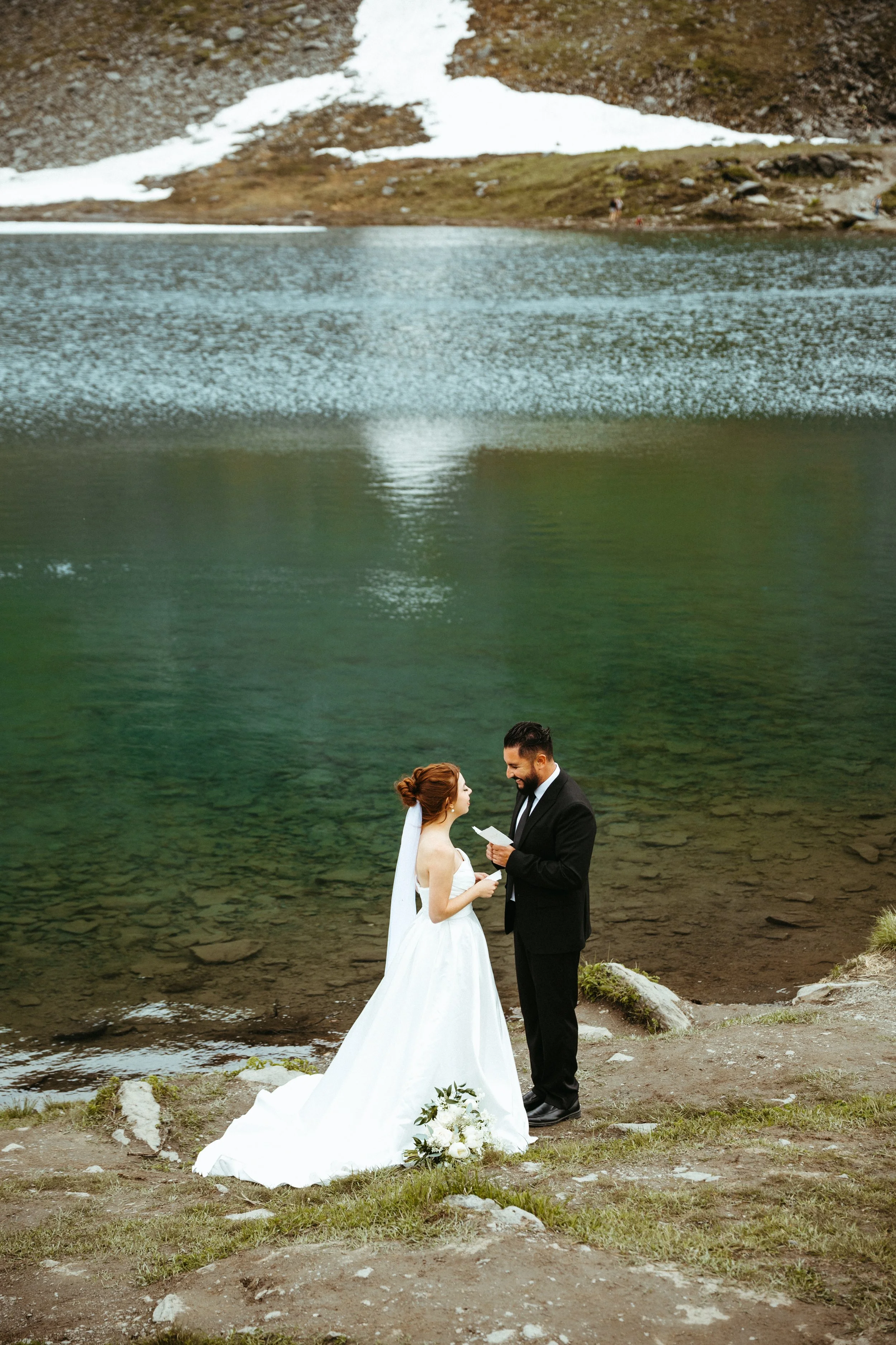 A bride and groom standing by a lake, with the groom reading vows from a paper during their wedding ceremony in a scenic mountain setting.