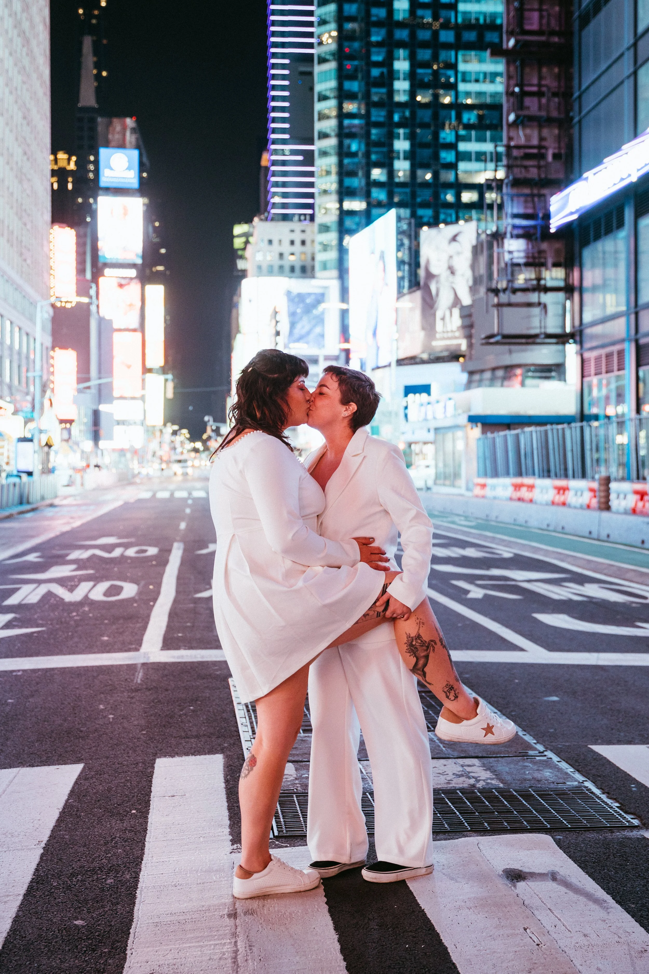 Two women wearing white outfits kissing in the middle of a city street at night with illuminated buildings and billboards in the background.