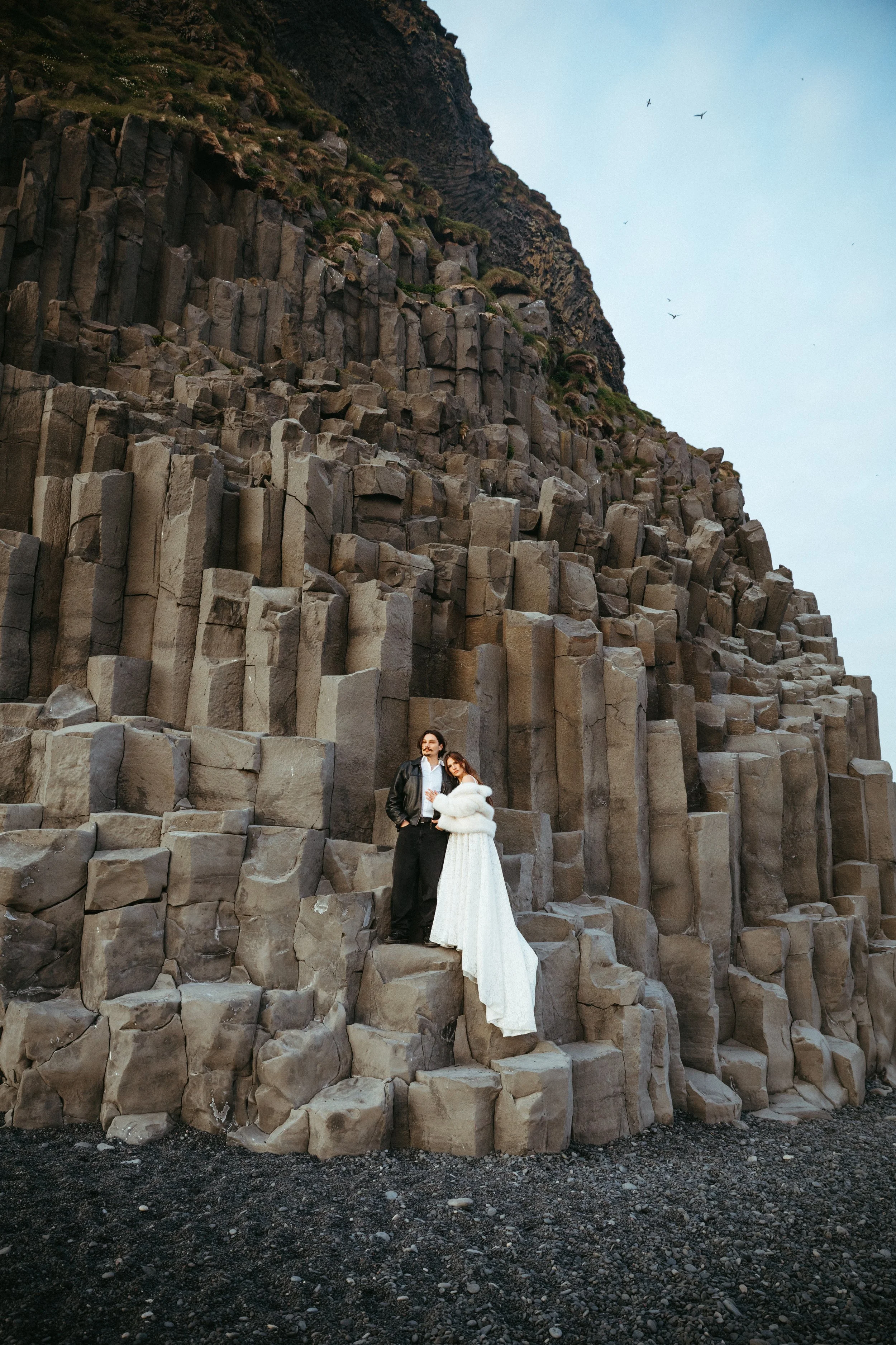 A couple in formal attire standing on basalt columns at the beach, with a cliff and sky in the background.