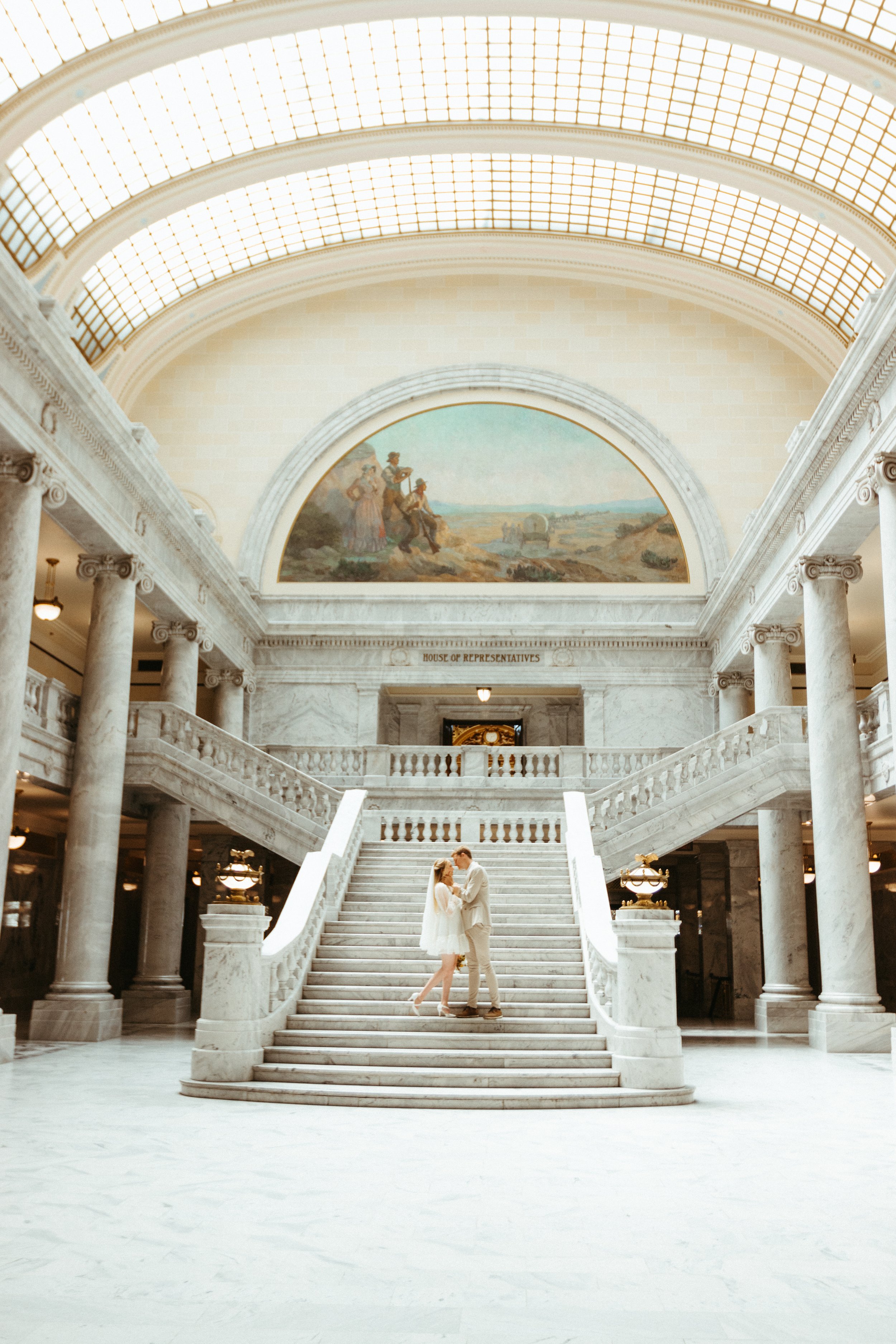 A couple in wedding attire standing on a grand marble staircase inside a historic building, with a mural and glass ceiling above.