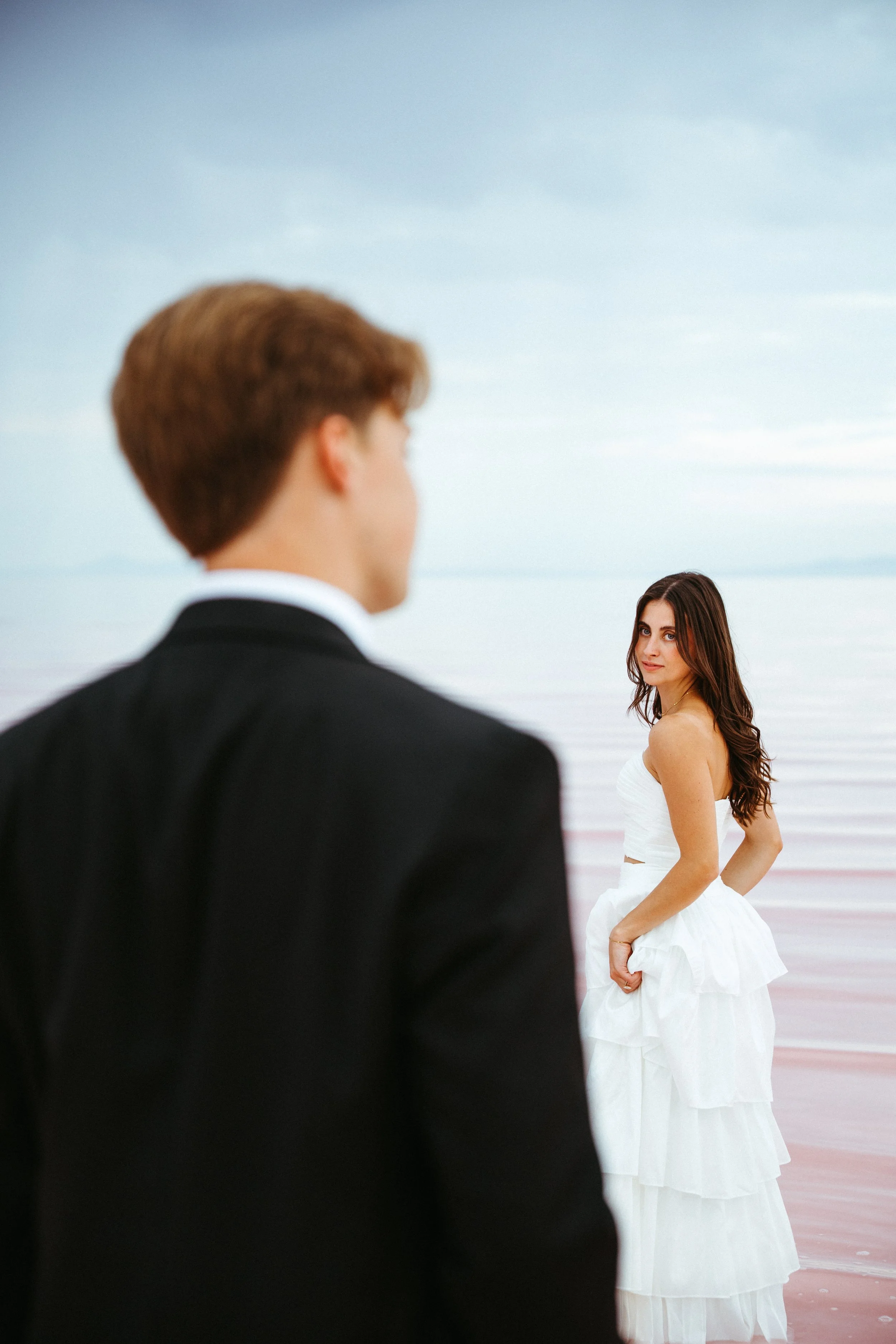 A man in a black suit with brown hair facing away from the camera, looking at a woman in a white dress with long brown hair standing near a body of water, with a cloudy sky above.