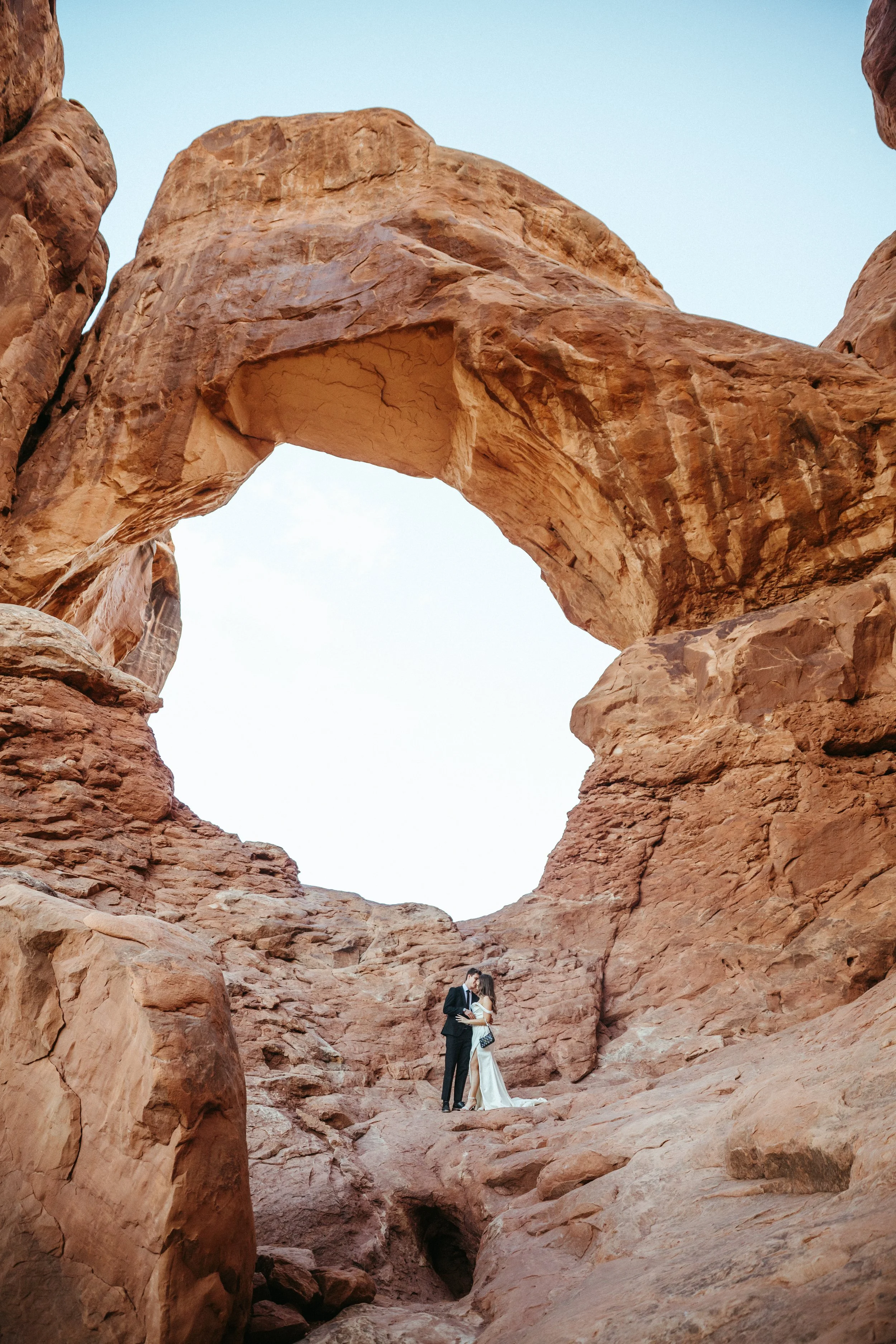 A couple dressed in wedding attire standing under a large natural rock arch in a desert landscape. 