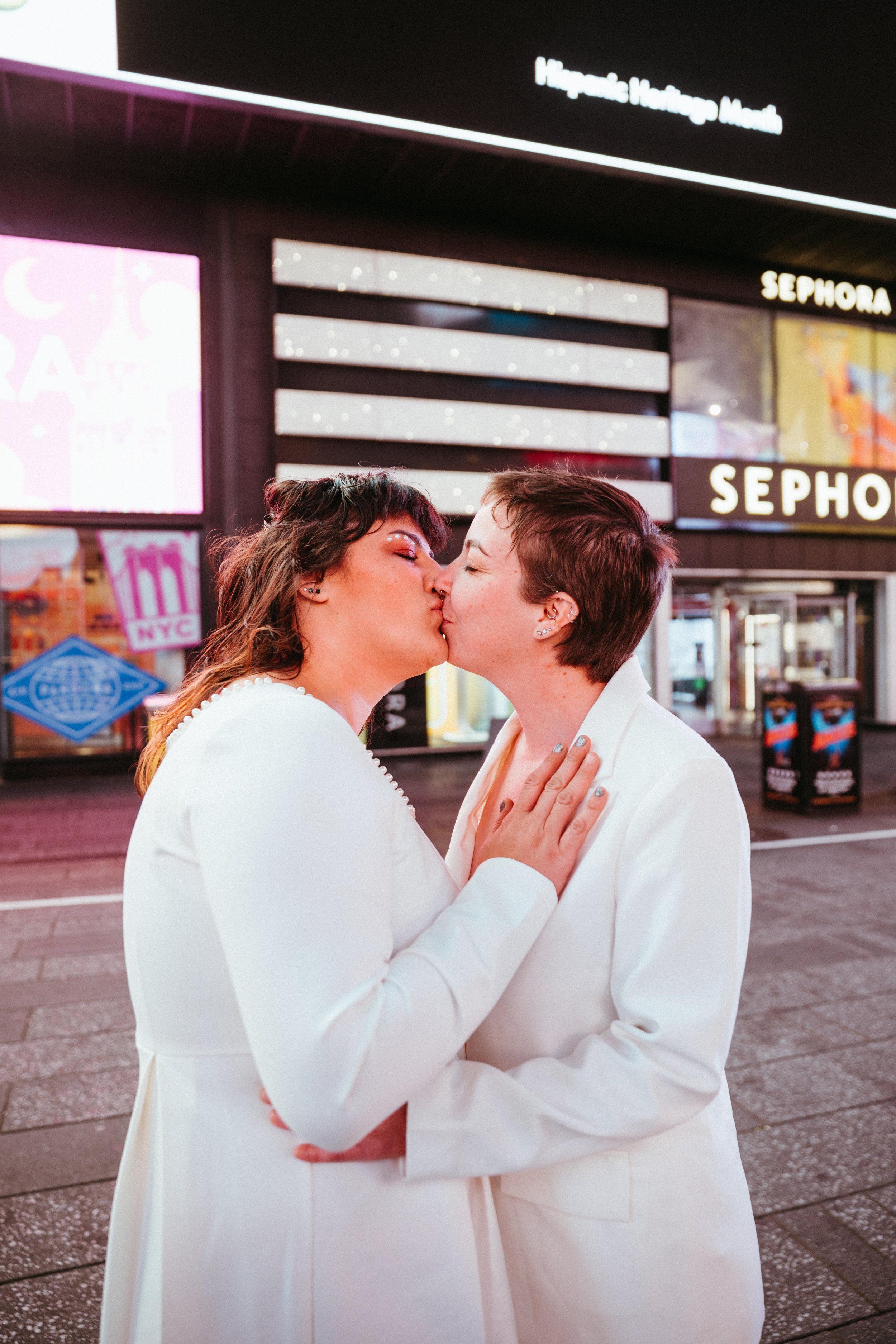 Two women in white dresses sharing a kiss in Times Square at night with bright lights and signage, including a Sephora store in the background.