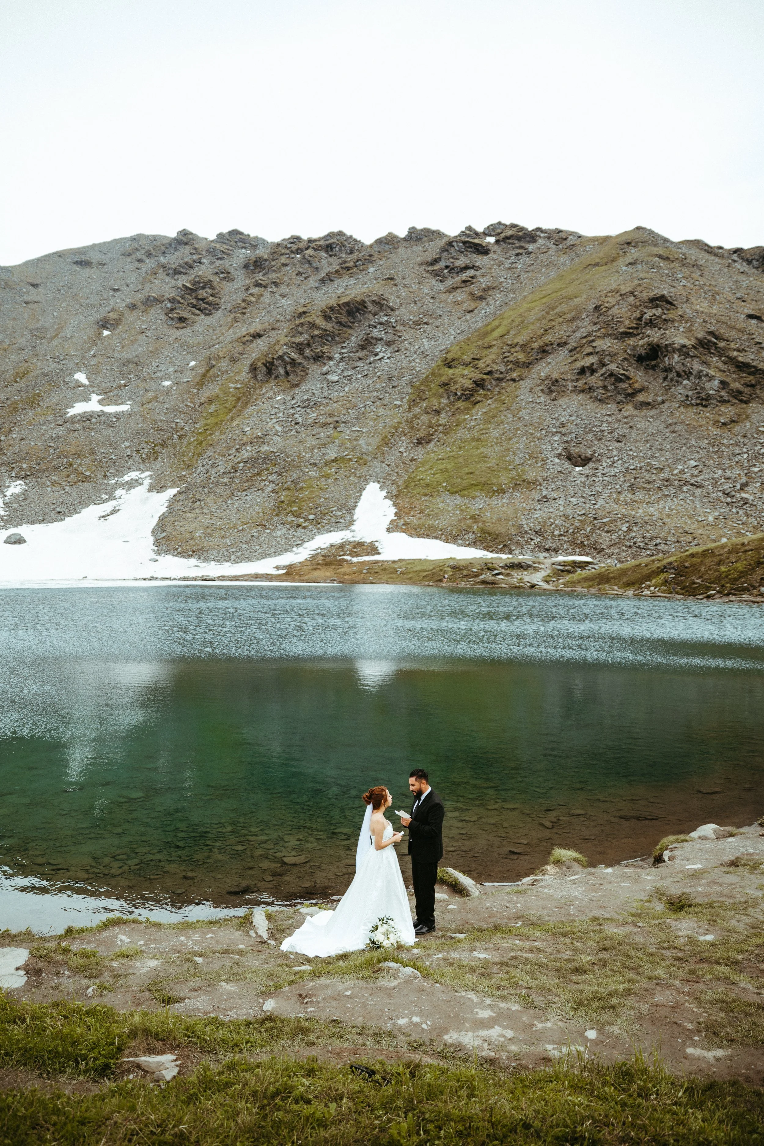 A bride and groom share a moment beside a mountain lake with rocky slopes and patches of snow.