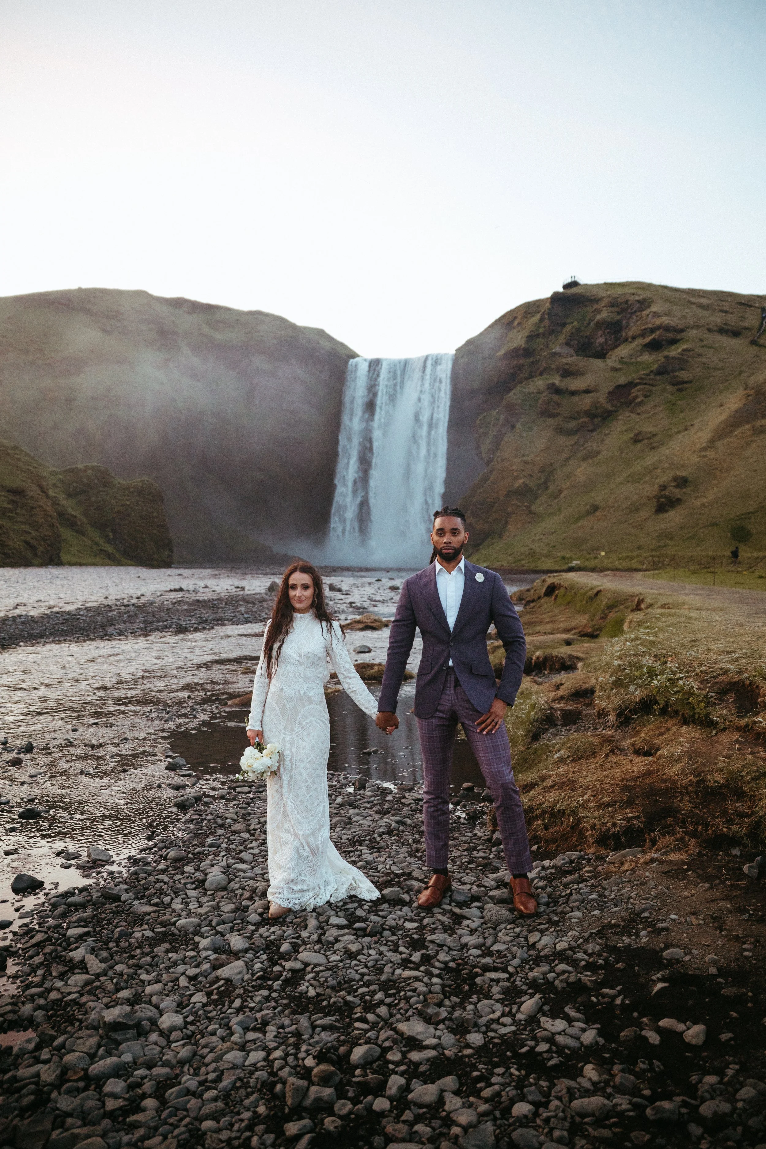A bride and groom standing hand-in-hand near a river with a waterfall in the background.