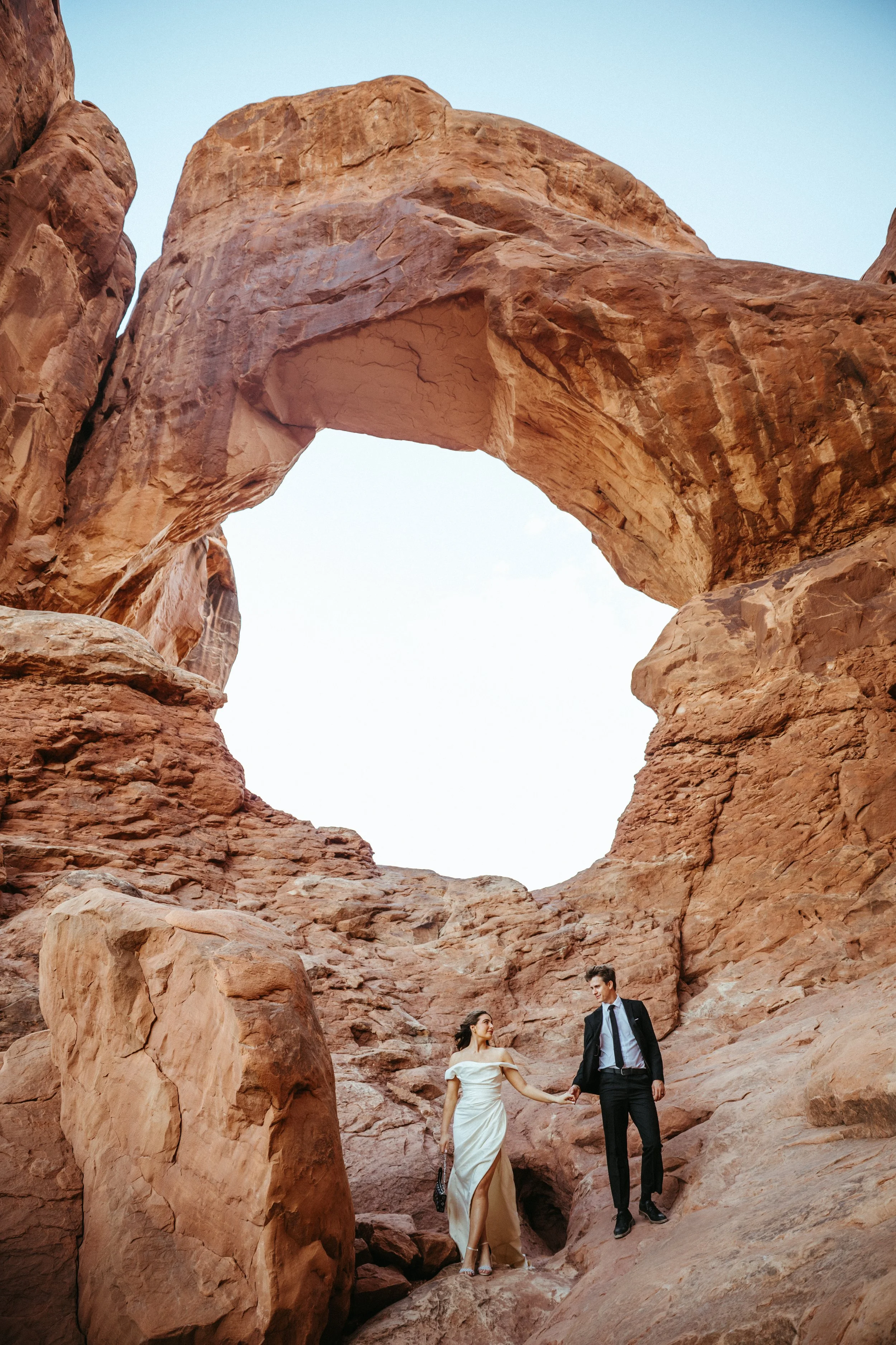 A man and woman holding hands, walking through large red rocks with a natural arch above them, in a desert landscape. Arches National Park wedding photographer, Moab Utah elopement photographer, southern Utah wedding photographer, adventure elopement