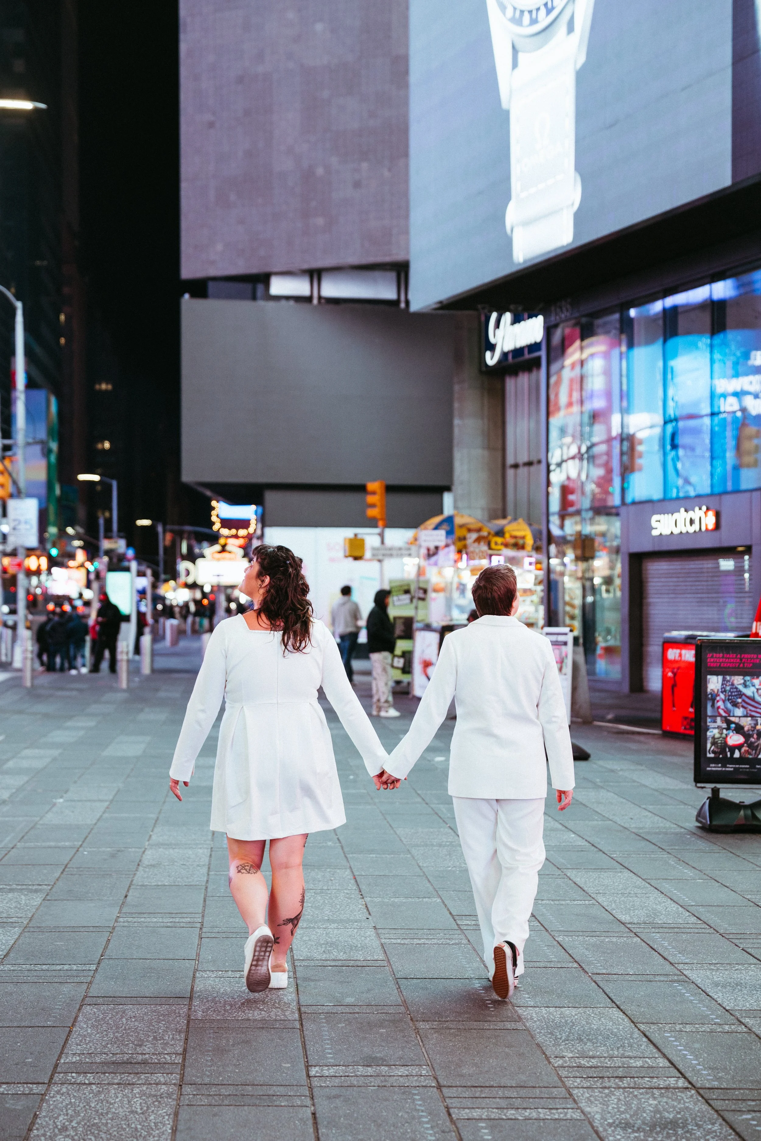 A couple holding hands walking in Times Square, New York City, at night, with bright billboards and people around.