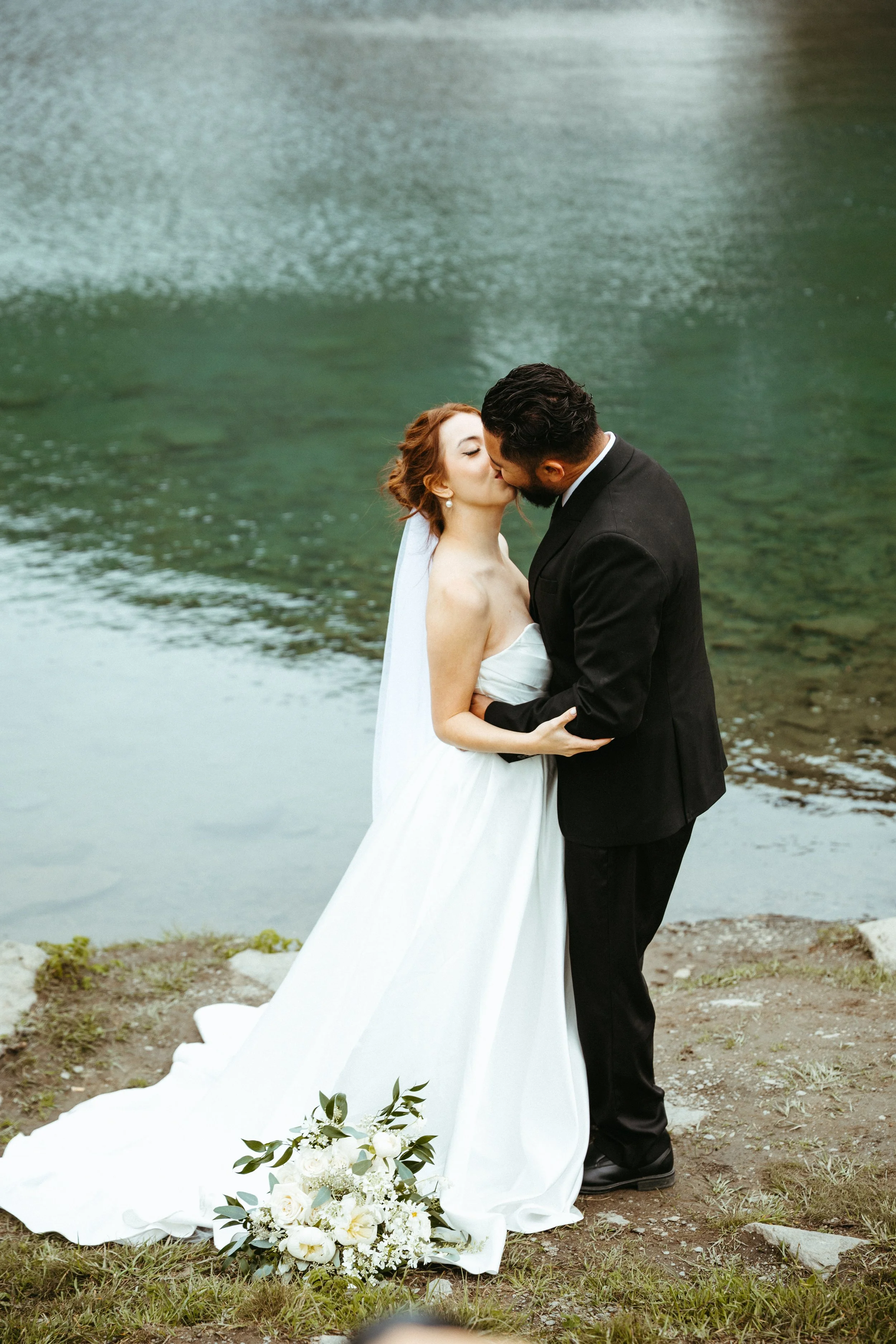 A bride and groom sharing a kiss outdoors by a river, with a bouquet of white flowers on the ground in front of them.