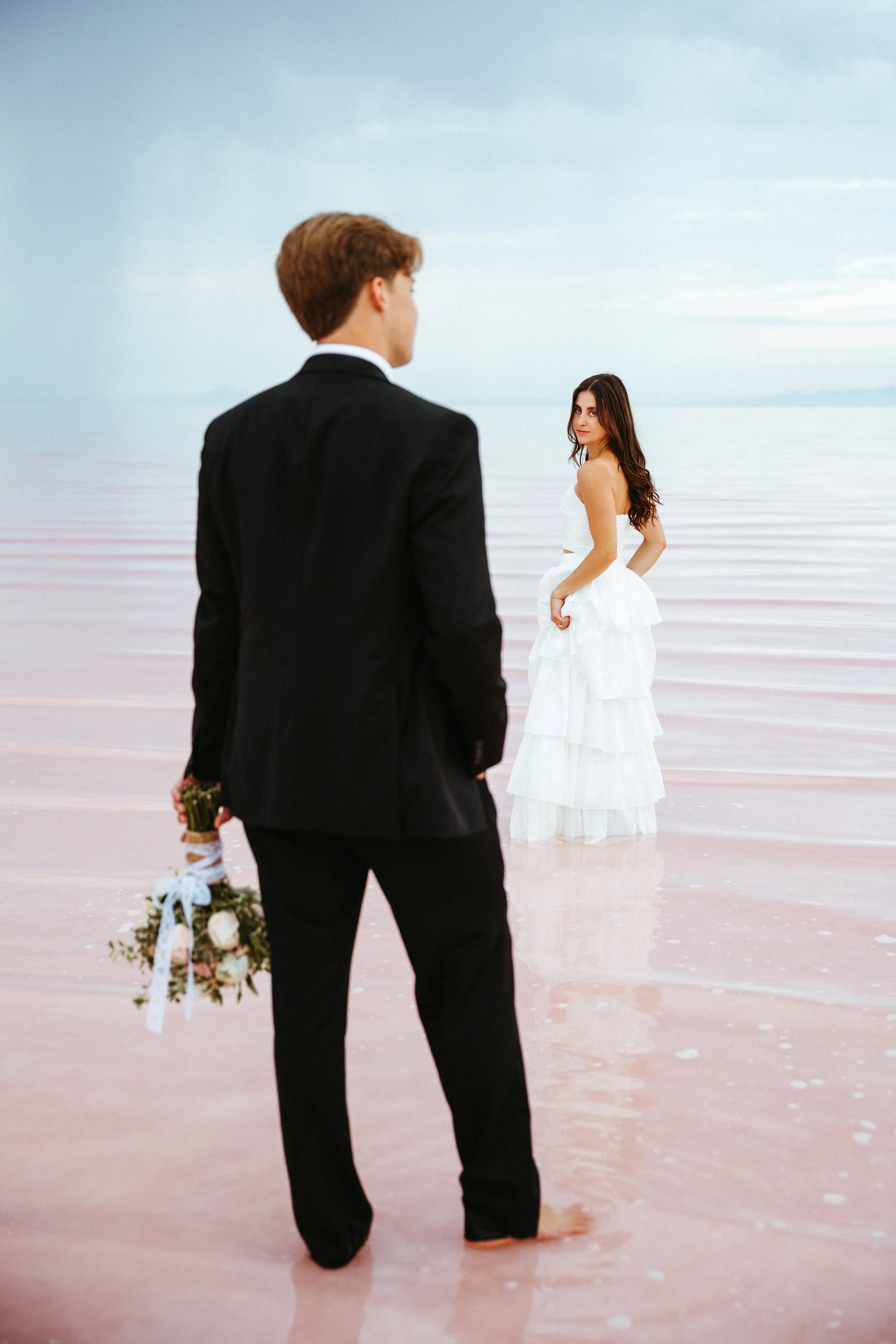A man in a black suit holding a bouquet of flowers stands in shallow water, facing a woman in a white dress standing in the water, with the ocean and sky in the background.