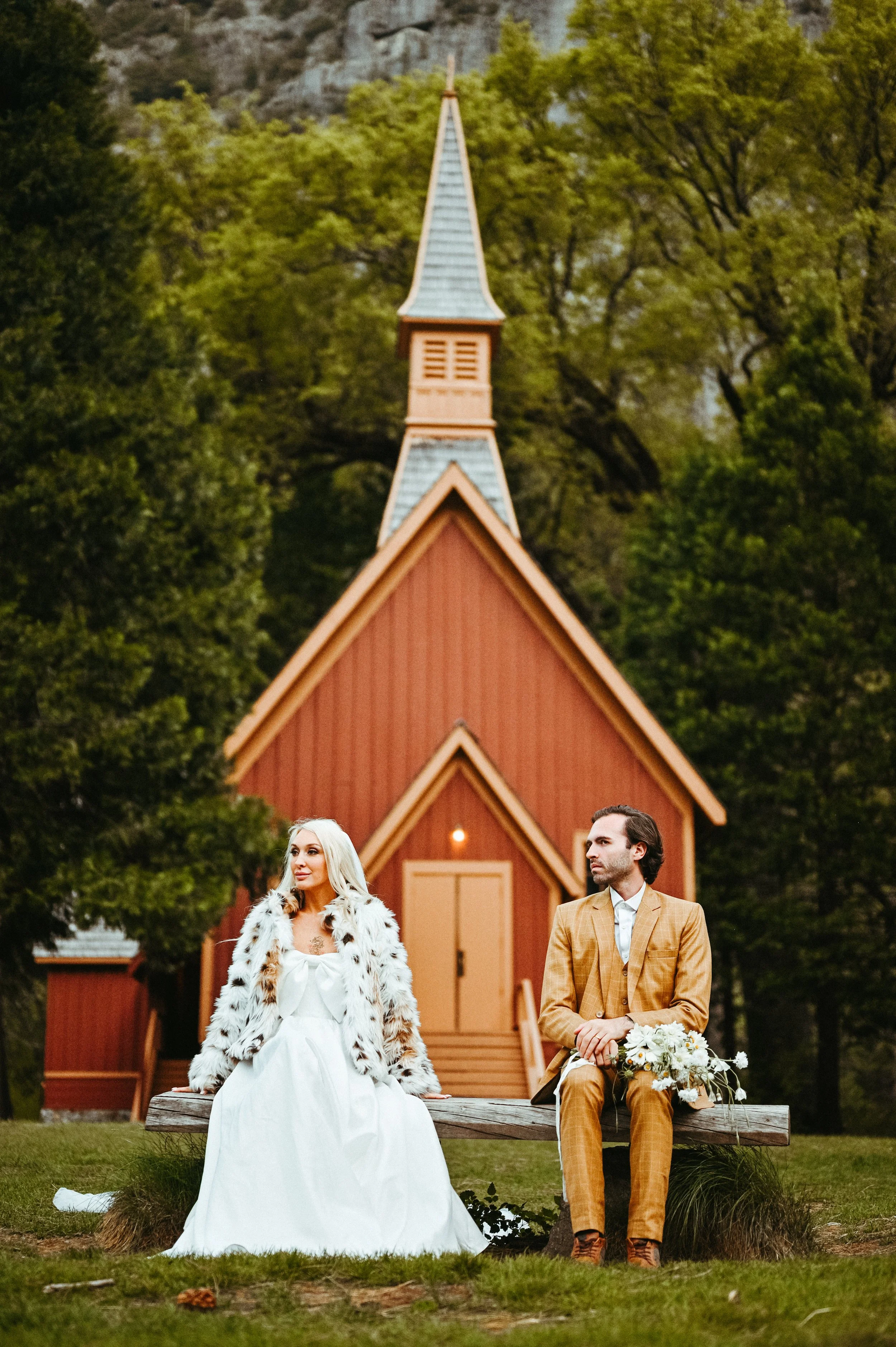 A woman in a white dress and faux fur coat and a man in a tan suit sitting on a wooden bench outdoors in front of a small red church with a tall steeple, surrounded by trees.