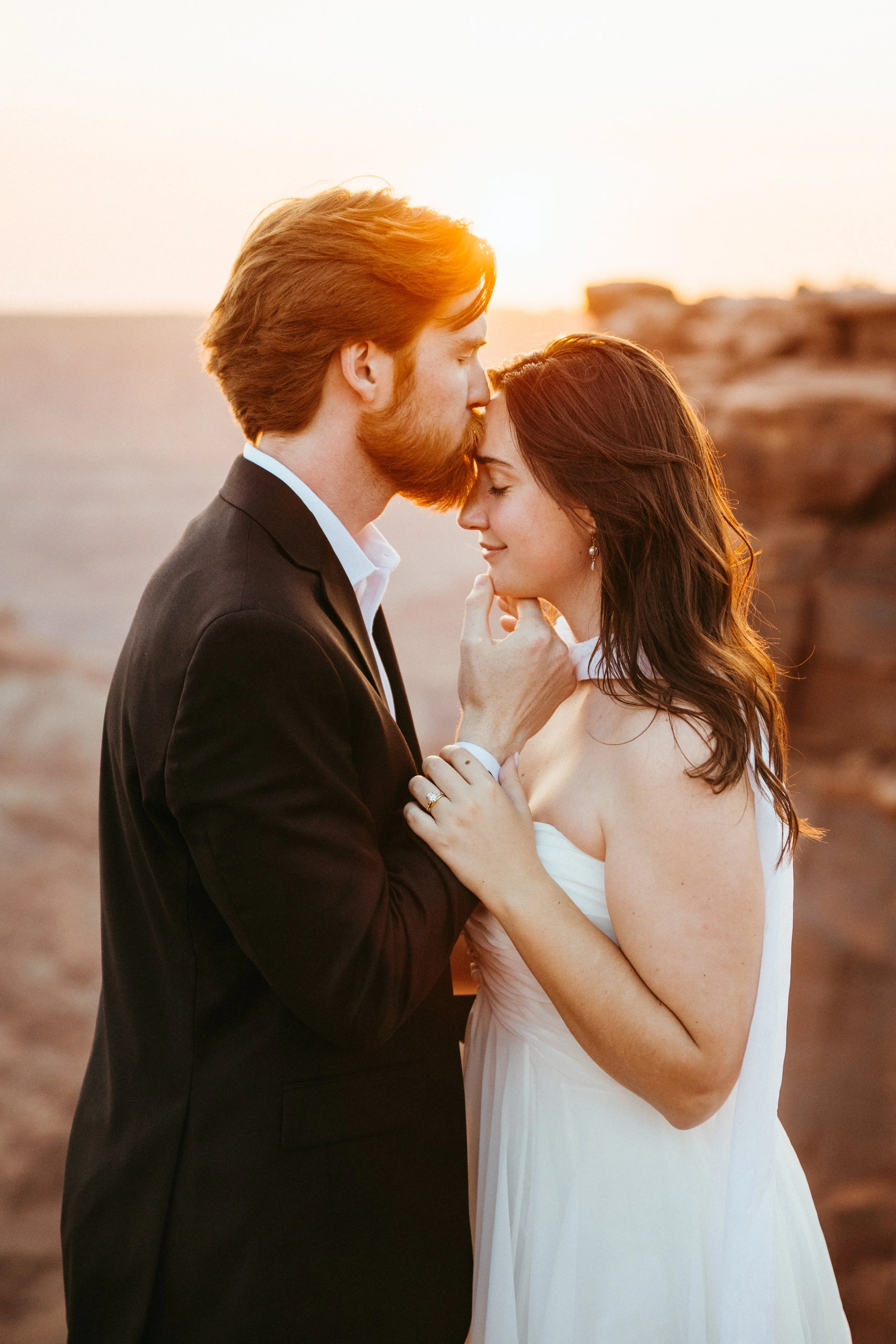 A couple, dressed in wedding attire, shares an intimate moment during sunset on a rocky coastline, with the man gently holding the woman's face and the woman smiling softly.