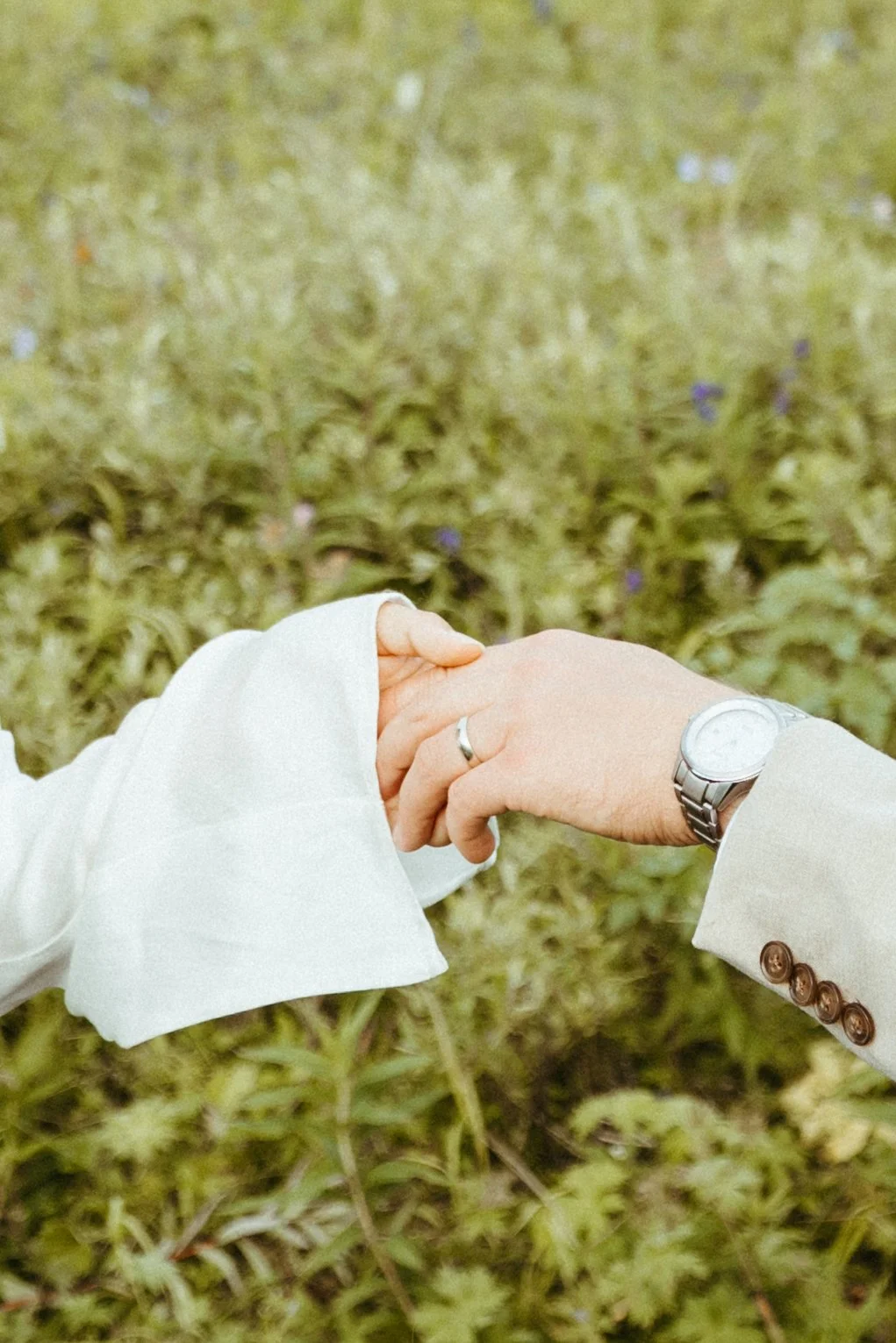 Two people holding hands outside amidst green grass and small purple flowers, one wearing a white long-sleeved shirt and the other wearing a beige blazer with a silver watch.