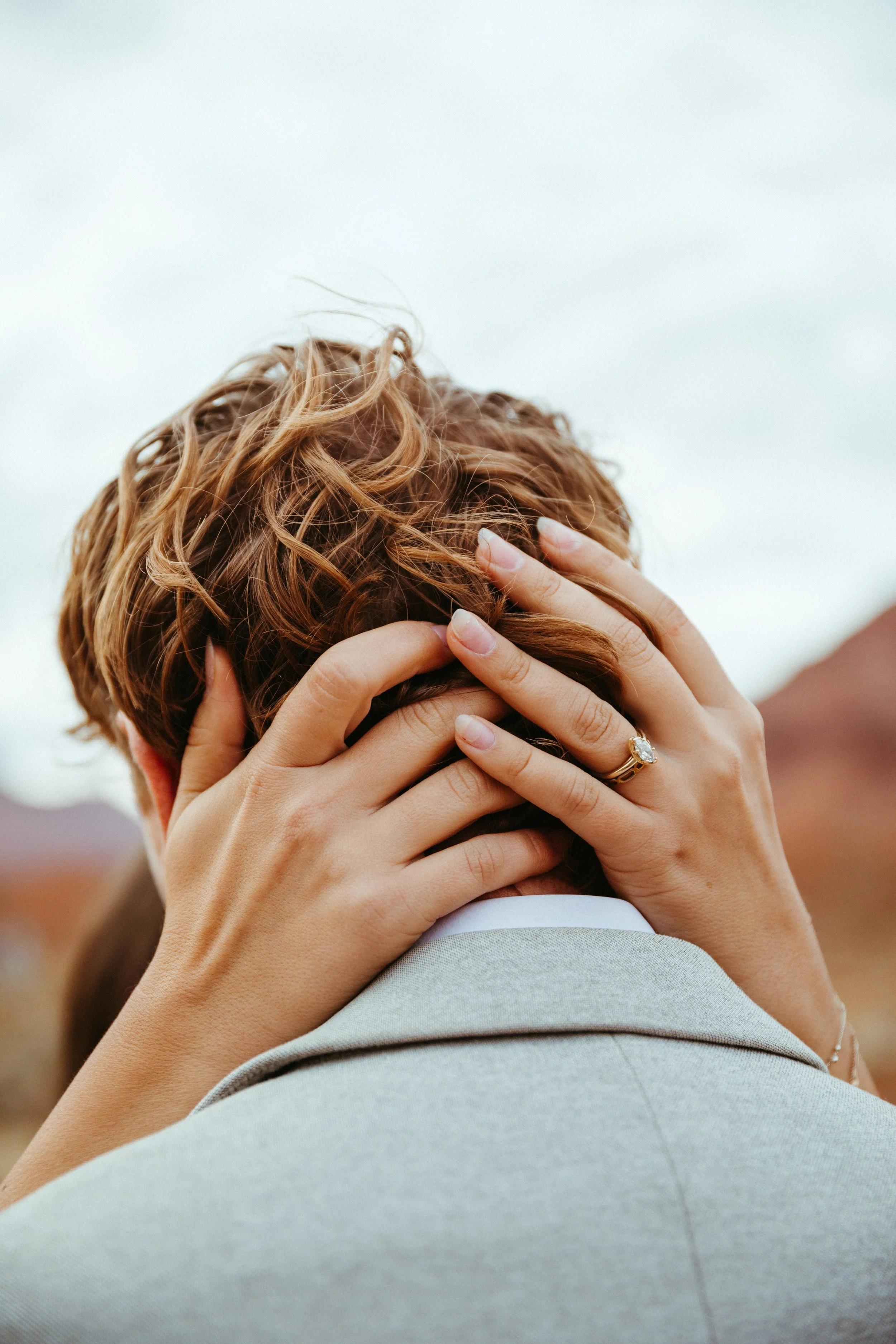 Close-up of a woman holding a man's head, showing her hand with an engagement ring, with woman's wavy hair and man's gray suit.