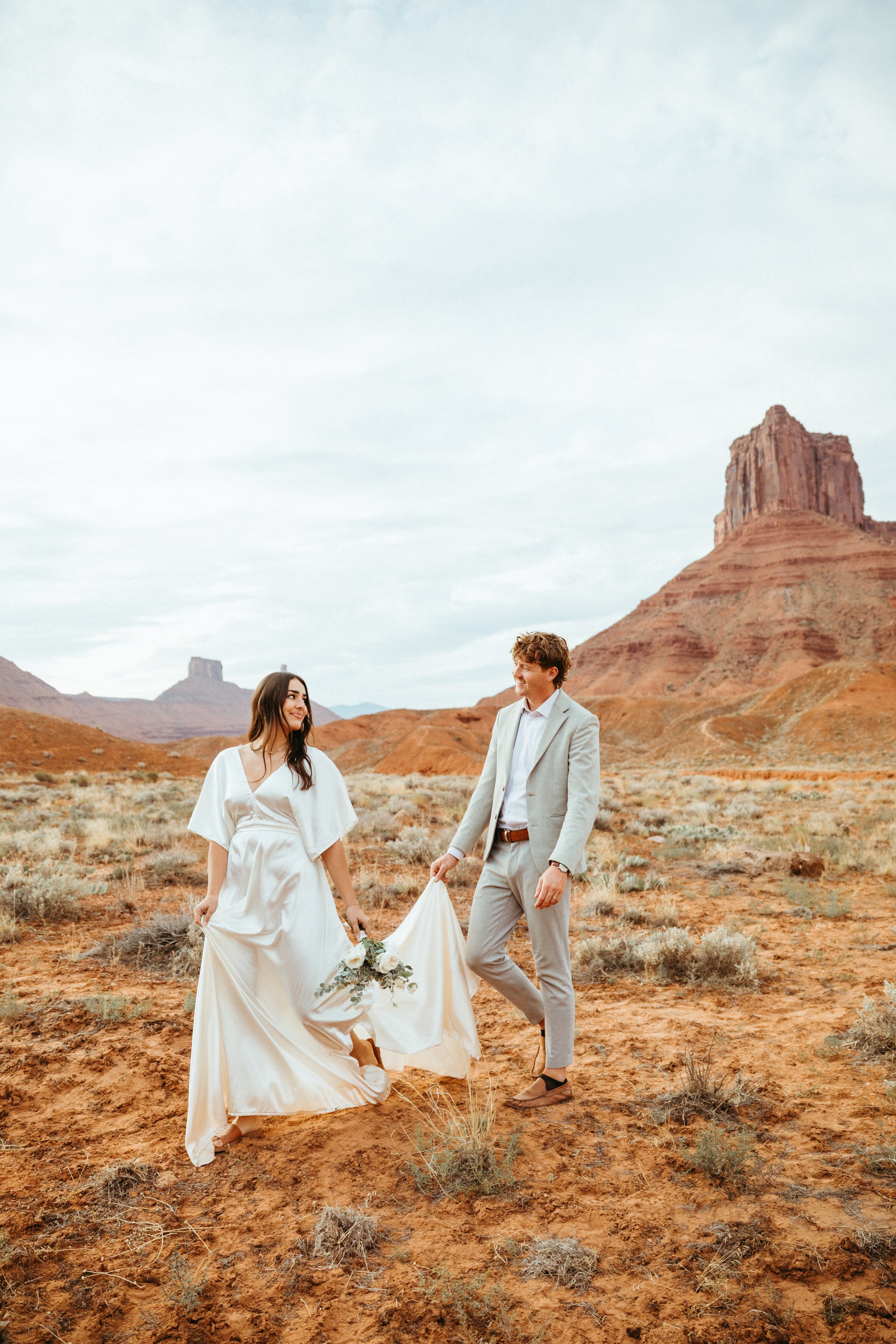 A bride and groom standing in a desert landscape with rock formations in the background, the bride wearing a white dress and holding a bouquet, and the groom in a light gray suit smiling at her.