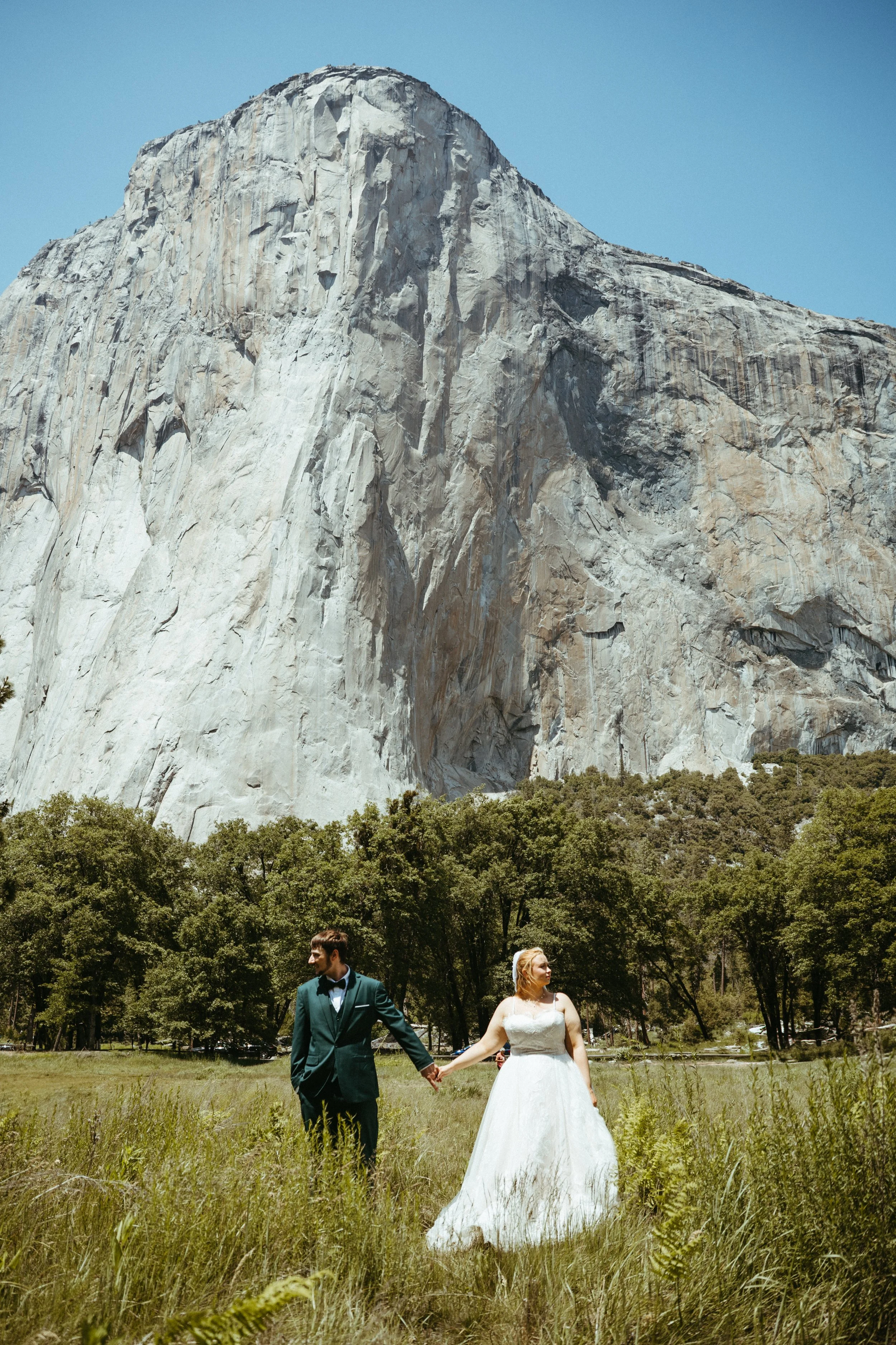 A bride and groom holding hands in a grassy field with trees and a large granite mountain in the background.
