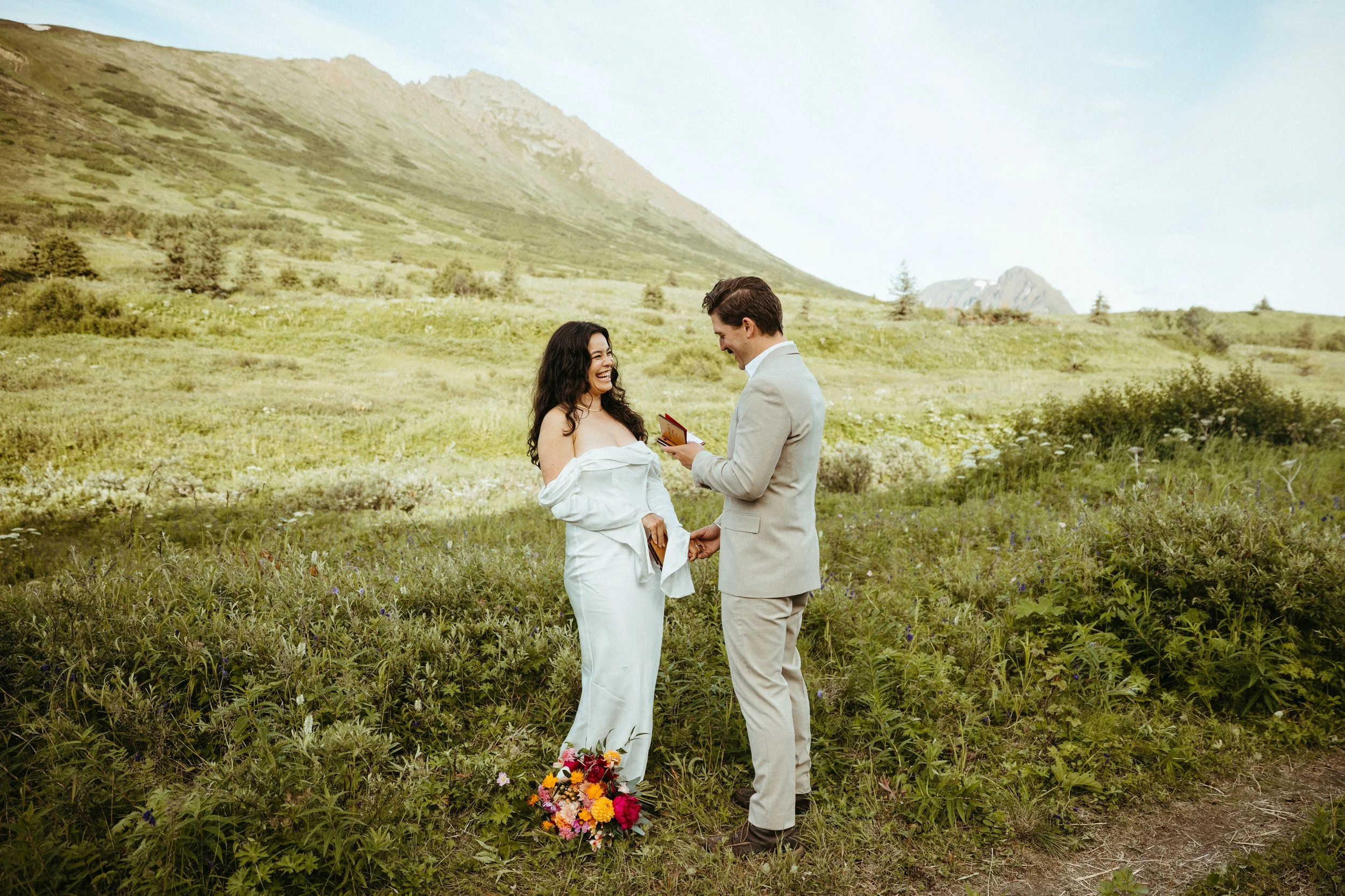 A couple getting married outdoors in a green field with mountains in the background. The woman is wearing a white dress and holding a bouquet of colorful flowers. The man is in a light suit, holding her hand, and reading from a small book. They are smiling at each other.