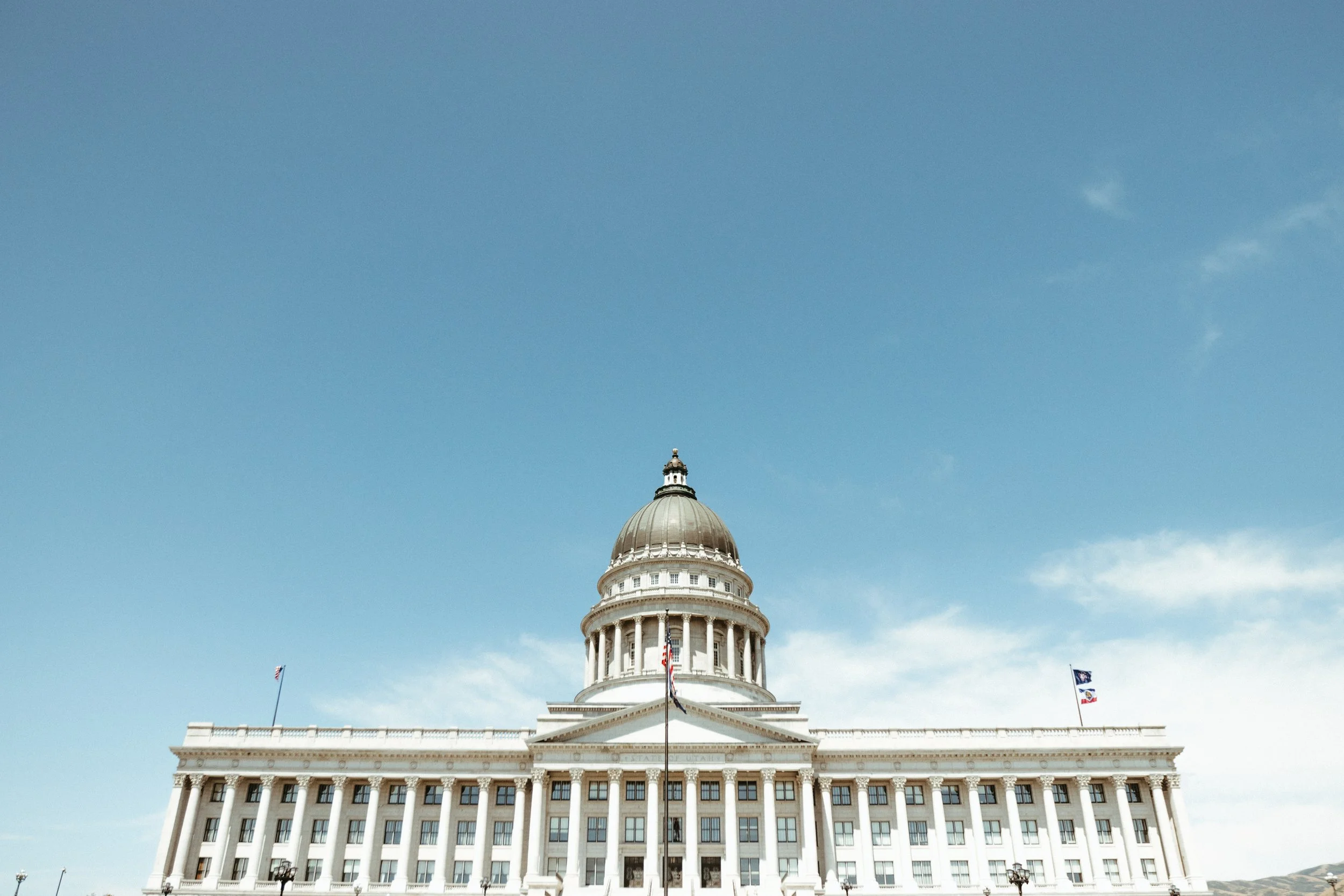 The image shows a large white government building with a prominent dome, columns, and flags on either side, set against a clear blue sky.