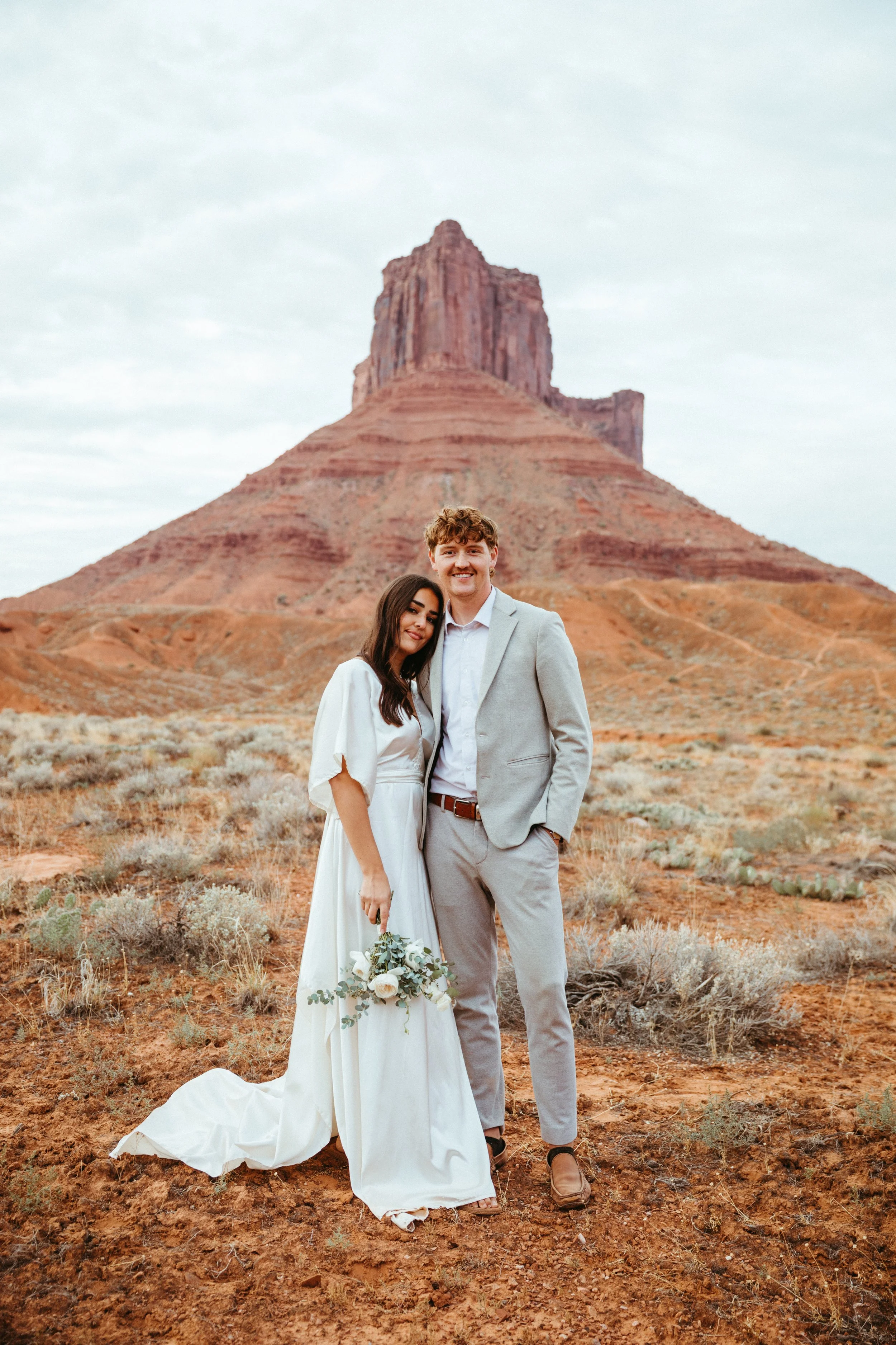 A newlywed couple standing in a desert landscape with a large red rock formation in the background, one woman in a white wedding dress holding a bouquet, and a man in a light gray suit smiling.