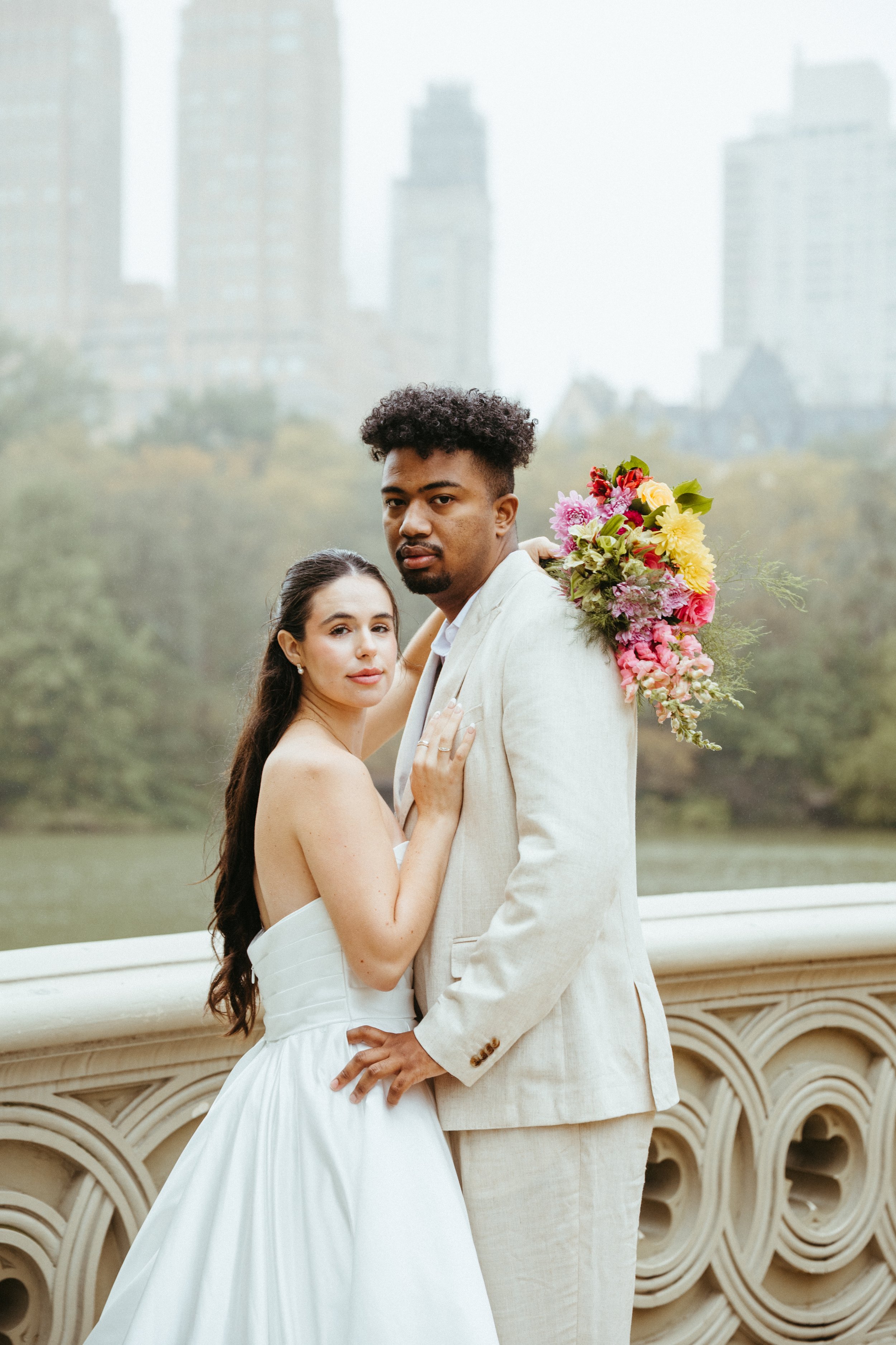 A couple dressed in wedding attire posing on a bridge with a city skyline in the background. The woman in a white wedding dress and the man in a light-colored suit, holding a colorful bouquet of flowers.