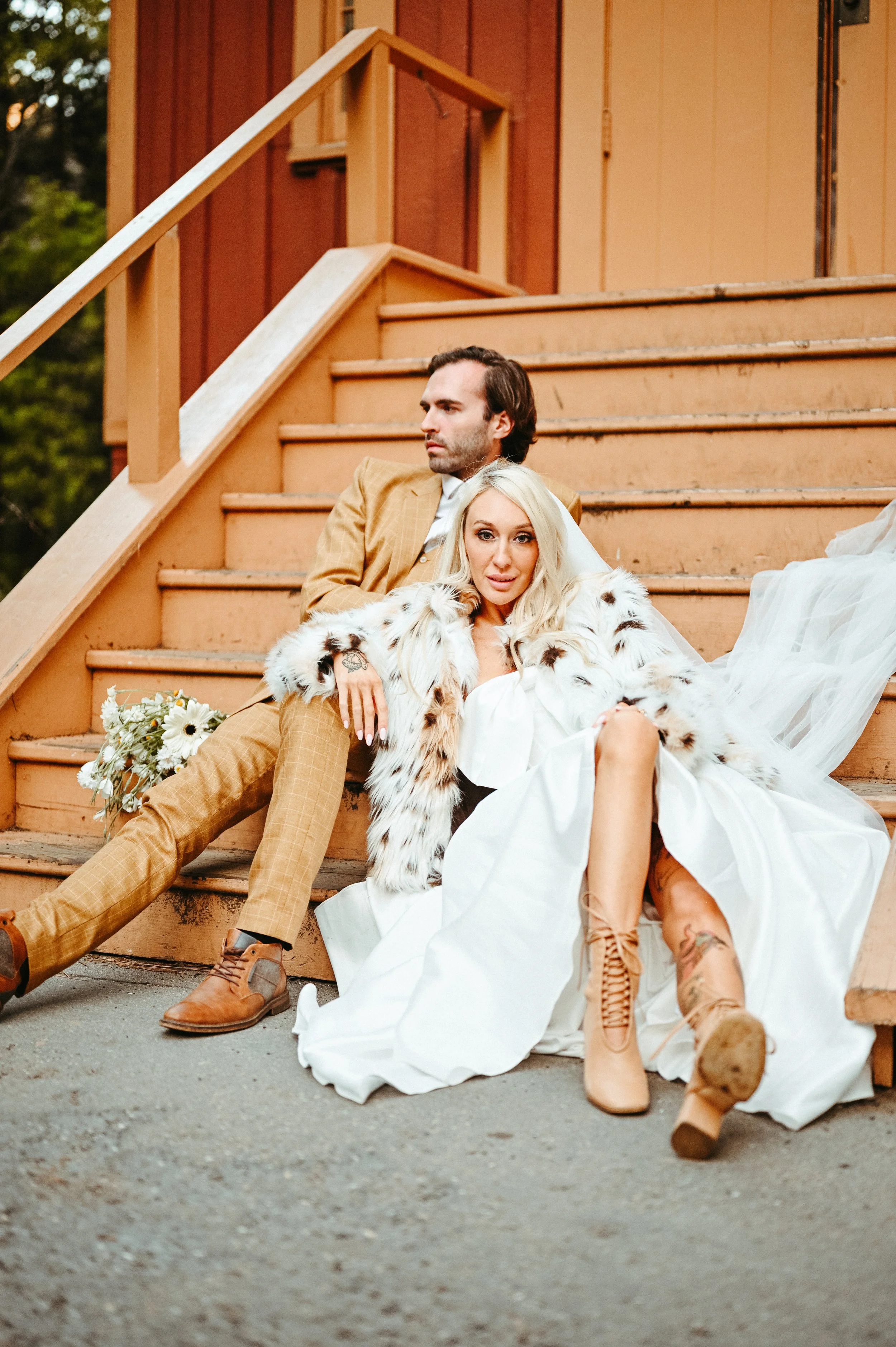 Couple sitting on wooden stairs, wearing wedding attire, with woman in a white dress and boots, man in plaid suit, woman with a fur coat, and bouquet of flowers nearby, outdoor setting.