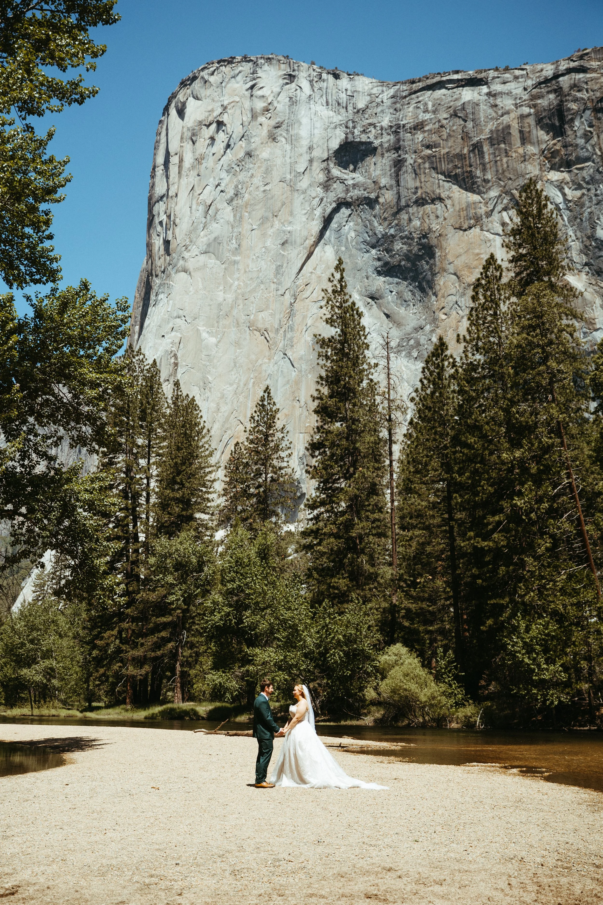 A bride in a white wedding dress and groom in a suit hold hands, standing by a river in a forested area with tall trees and a large rock formation in the background.