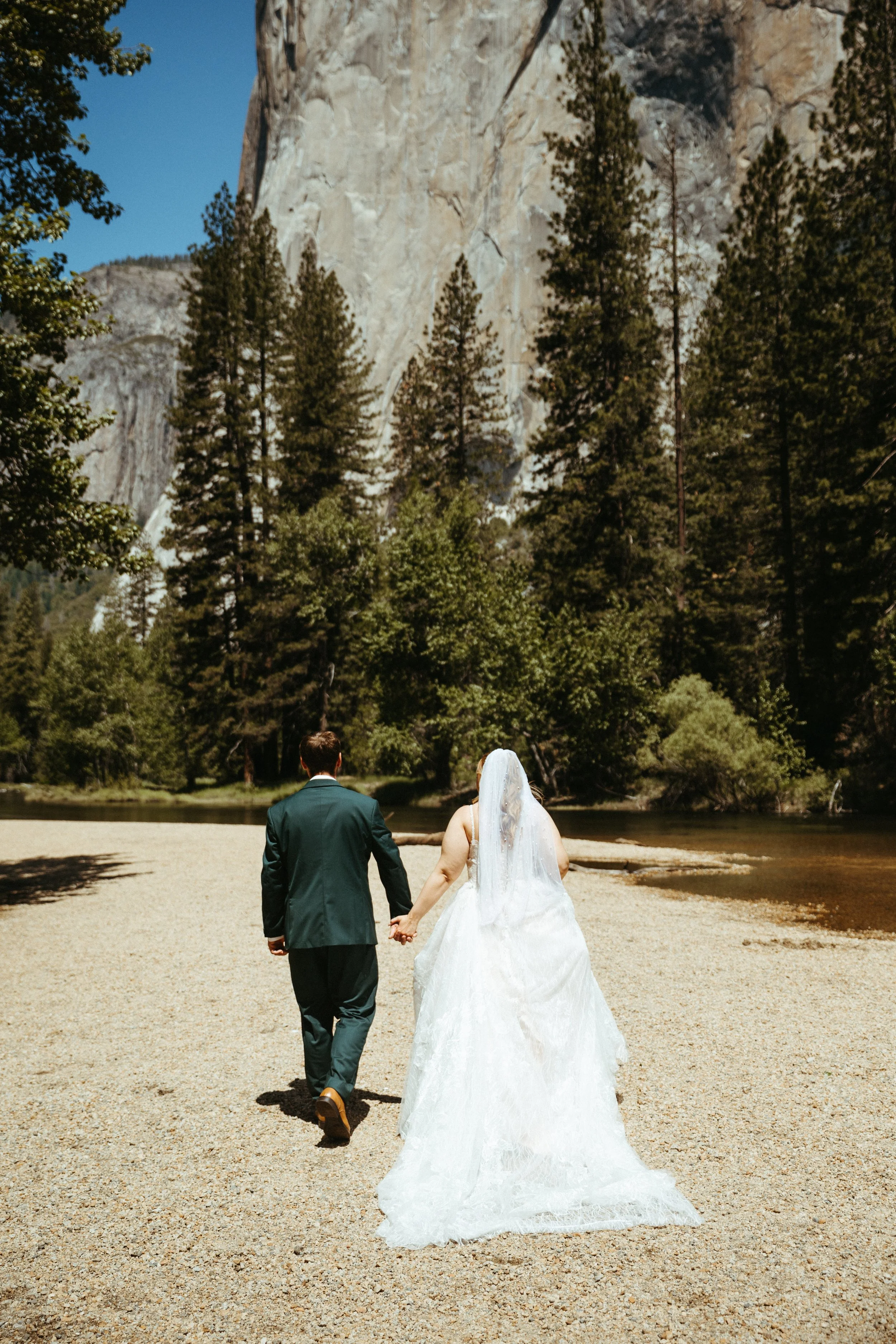 A bride and groom holding hands and walking away on a gravel path near a river, surrounded by trees and tall mountains in the background.