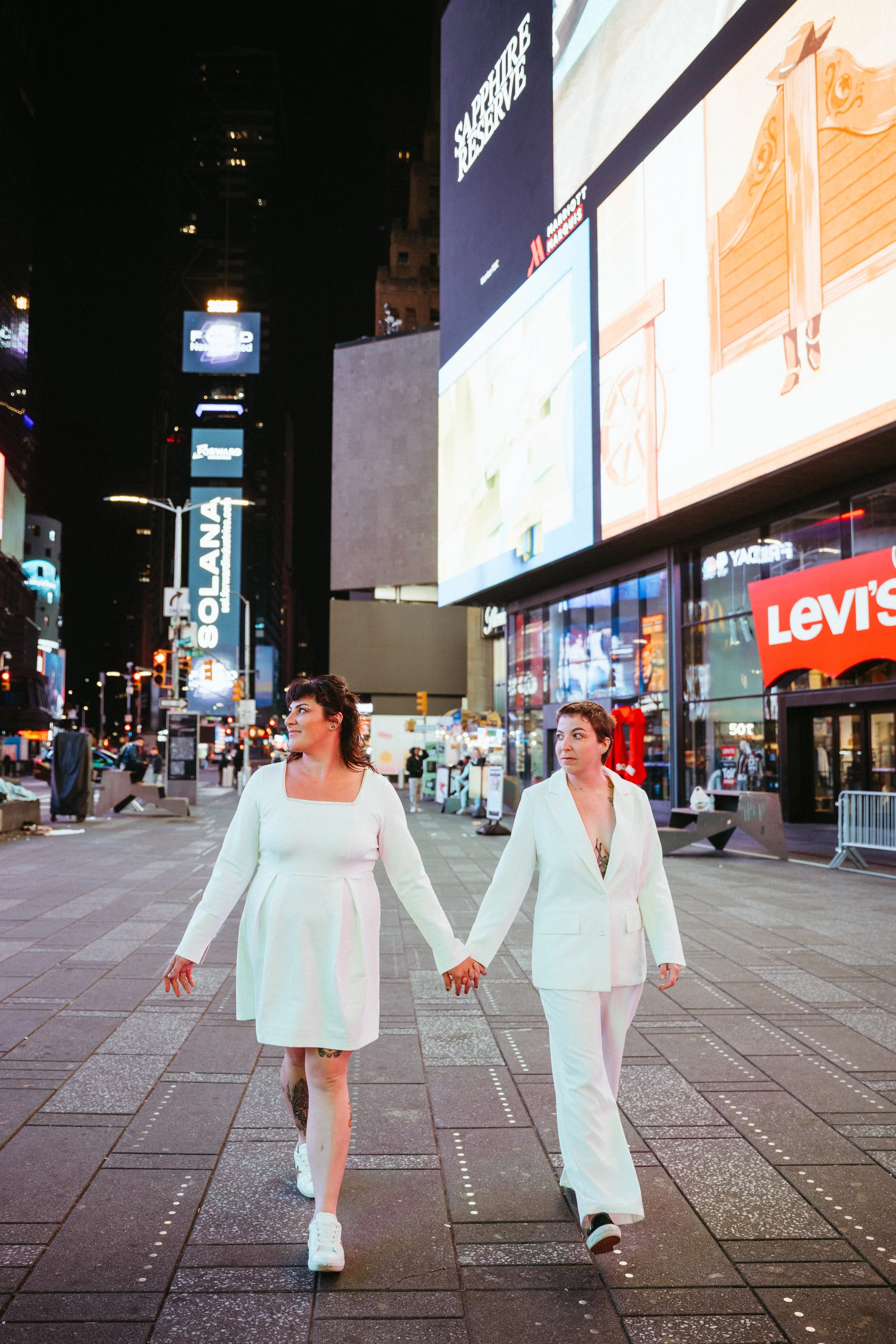 Two women in white outfits holding hands and walking in Times Square at night, surrounded by illuminated billboards and advertisements.