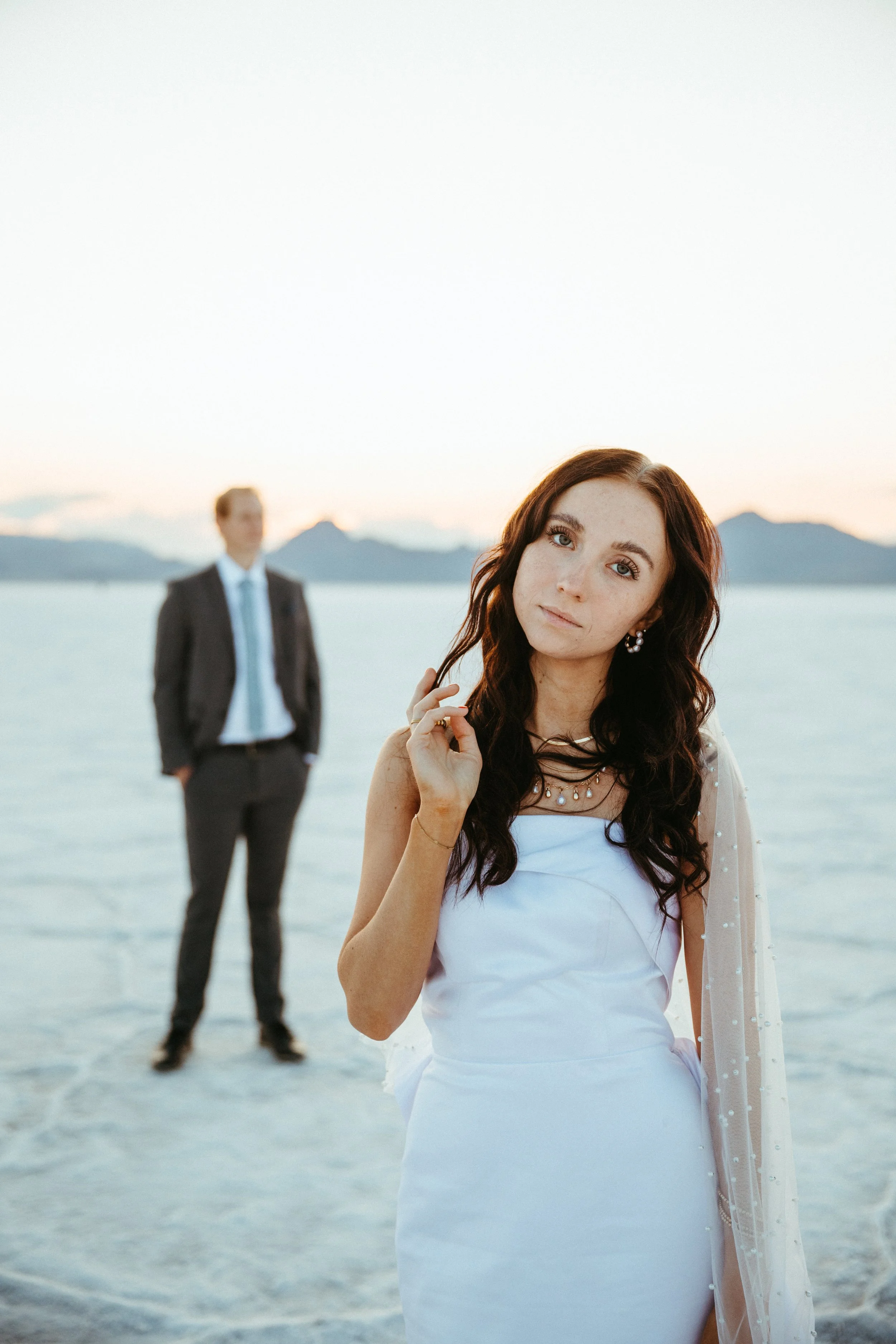 A woman in a white dress standing in front of a man in a suit on a salt flat at sunset with mountains in the background.