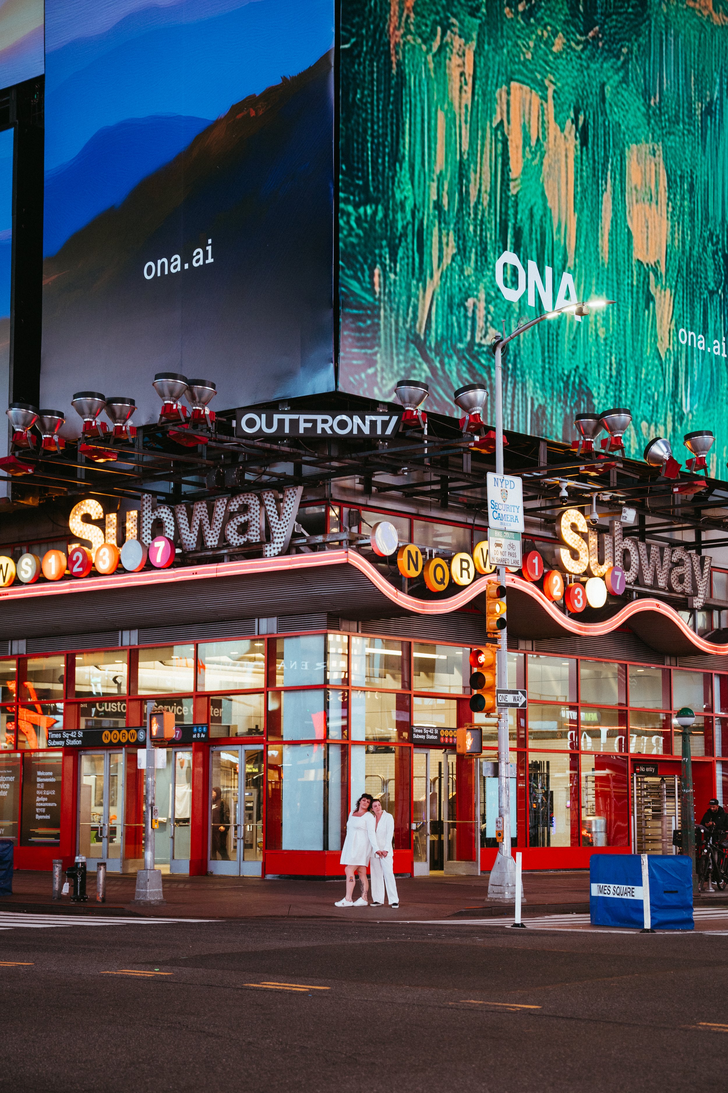 Nighttime scene at a NYC subway entrance with bright pink and red lights, large digital billboards displaying 'ona.ai', two women in white clothing standing in front, and traffic lights and signs on the street corner.