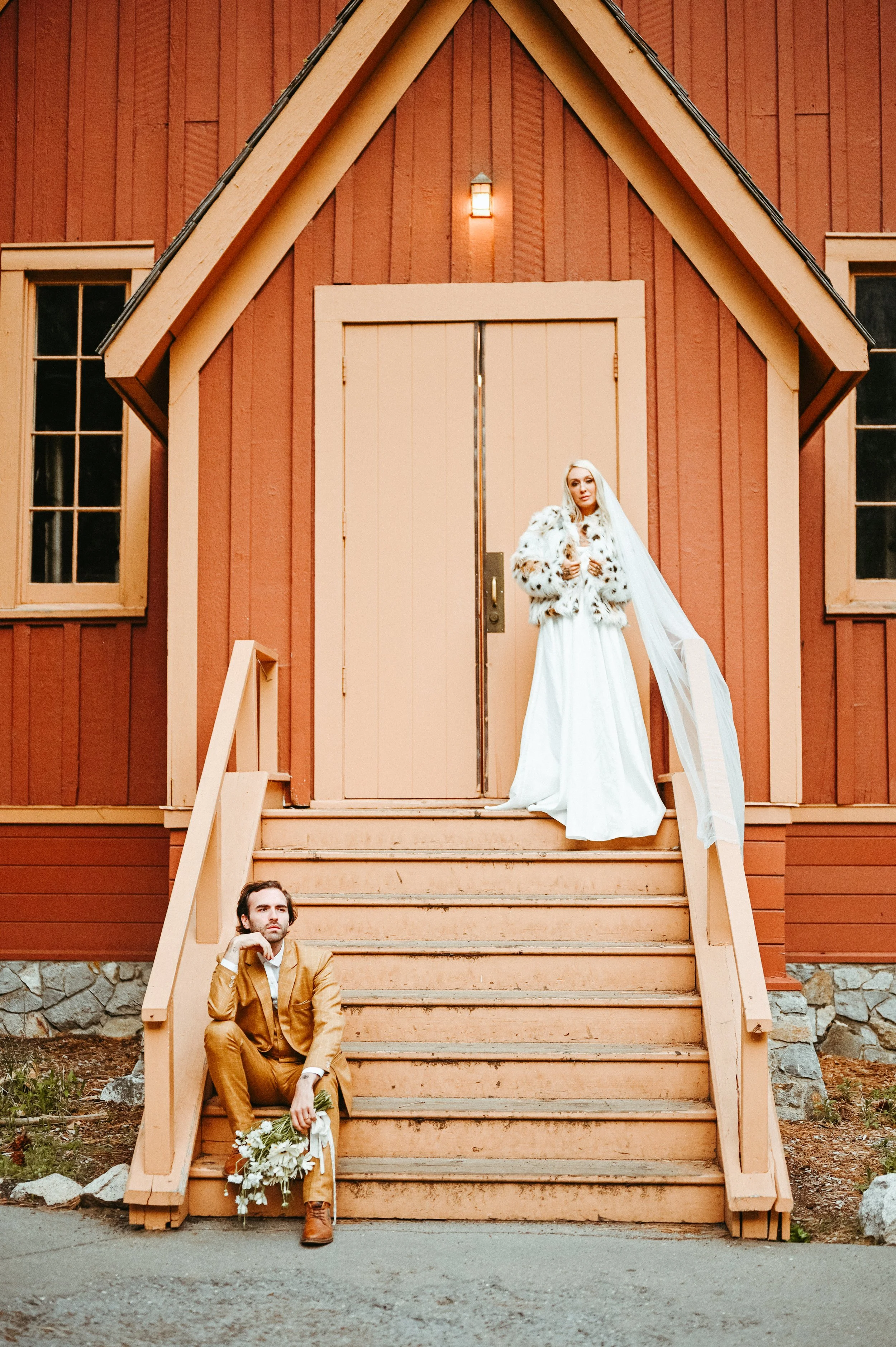 A bride and groom pose outside a rustic, orange wooden building, with the bride standing on the top of the stairs and the groom sitting on the steps below, holding a bouquet of flowers.