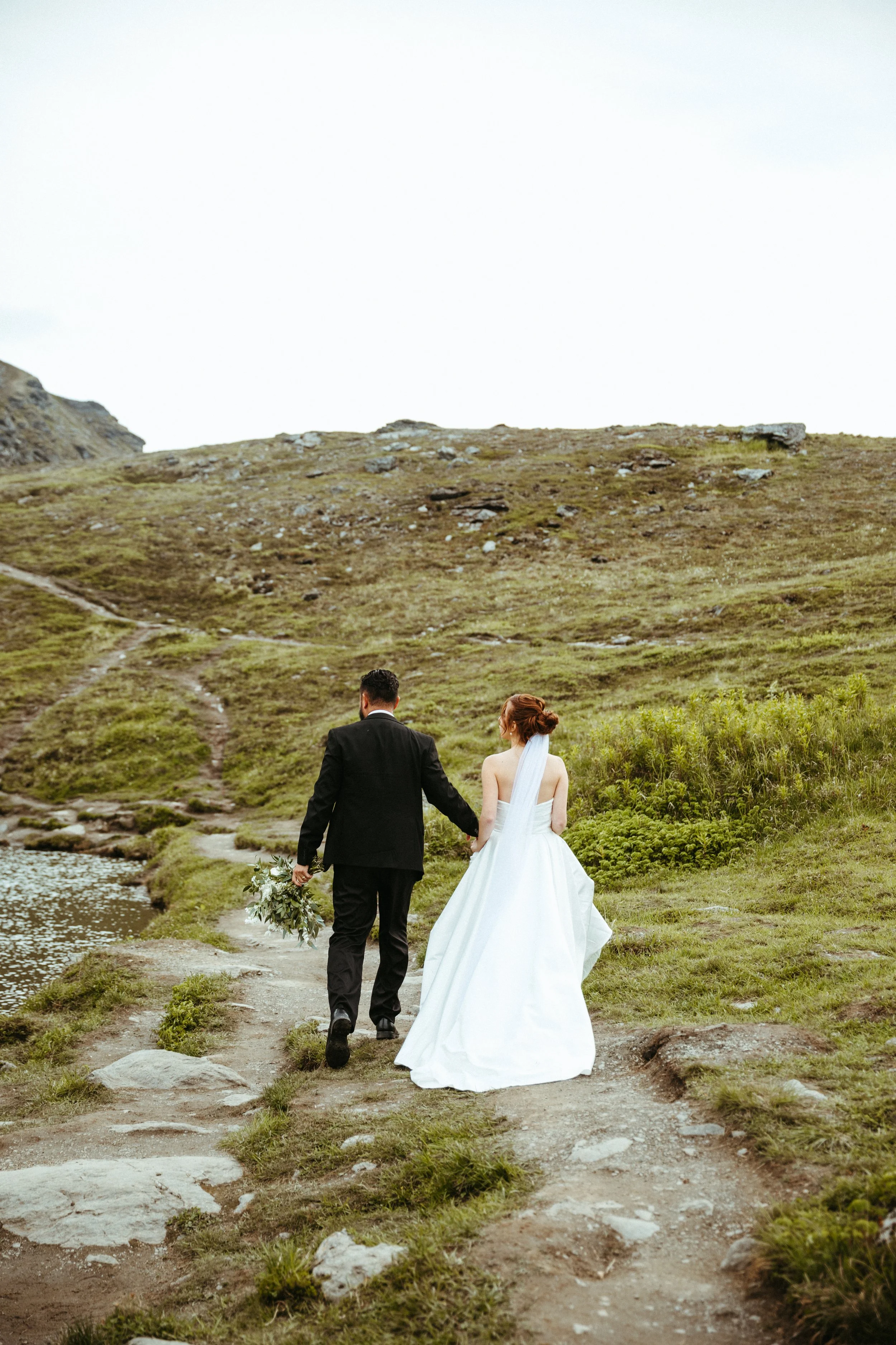 A bride and groom walking hand in hand along a dirt path beside a small body of water on a grassy hillside, with the bride in a white wedding dress and the groom in a black suit, holding a bouquet.