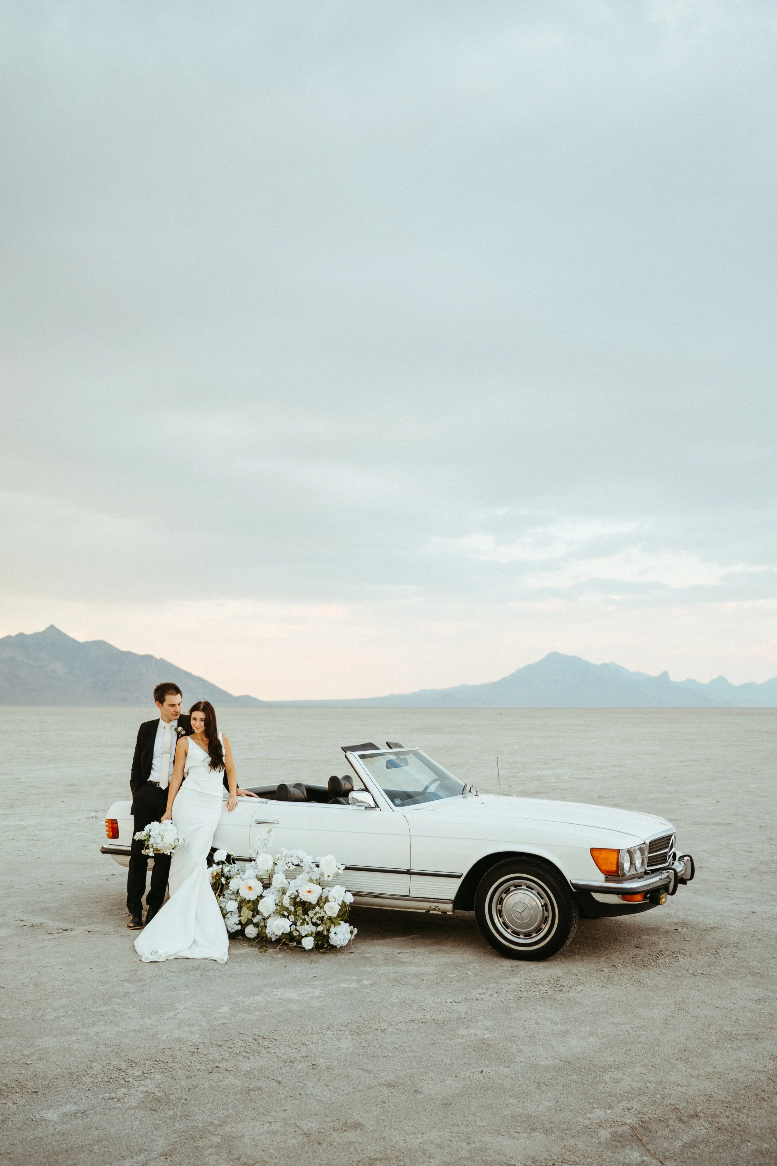 A newlywed couple in wedding attire standing next to a vintage white convertible car, decorated with white flowers, on a barren landscape with mountains and lake in the background.