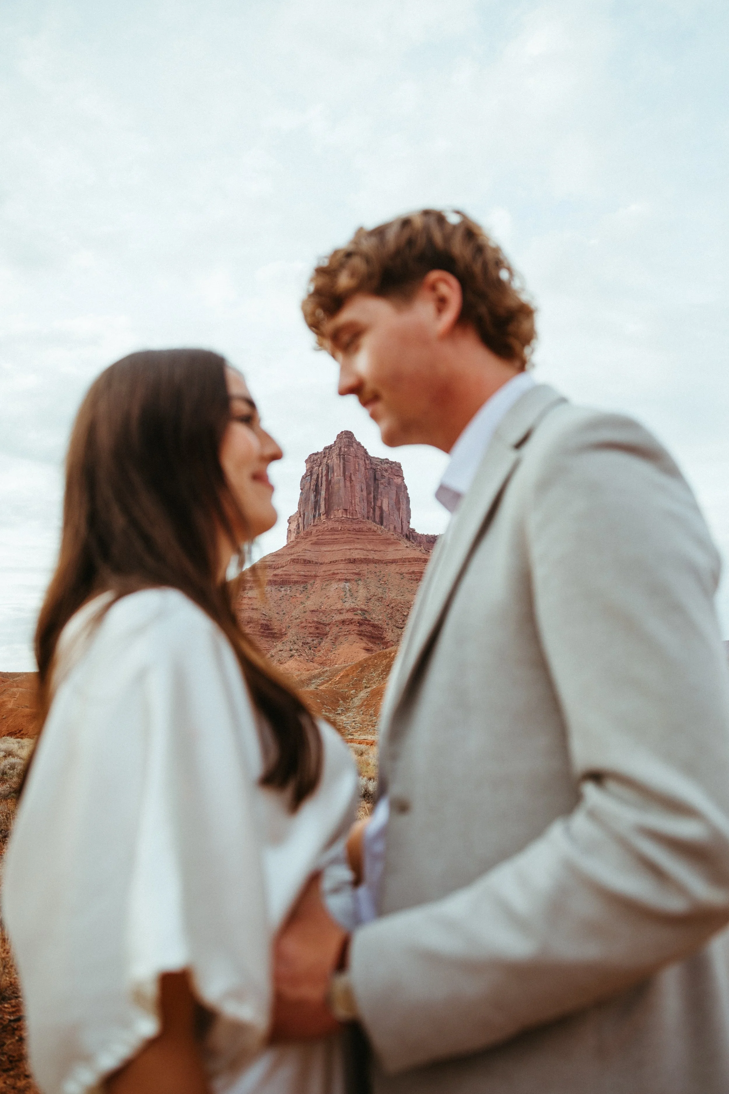 A couple holding hands and smiling at each other outdoors with a desert mountain landscape in the background.