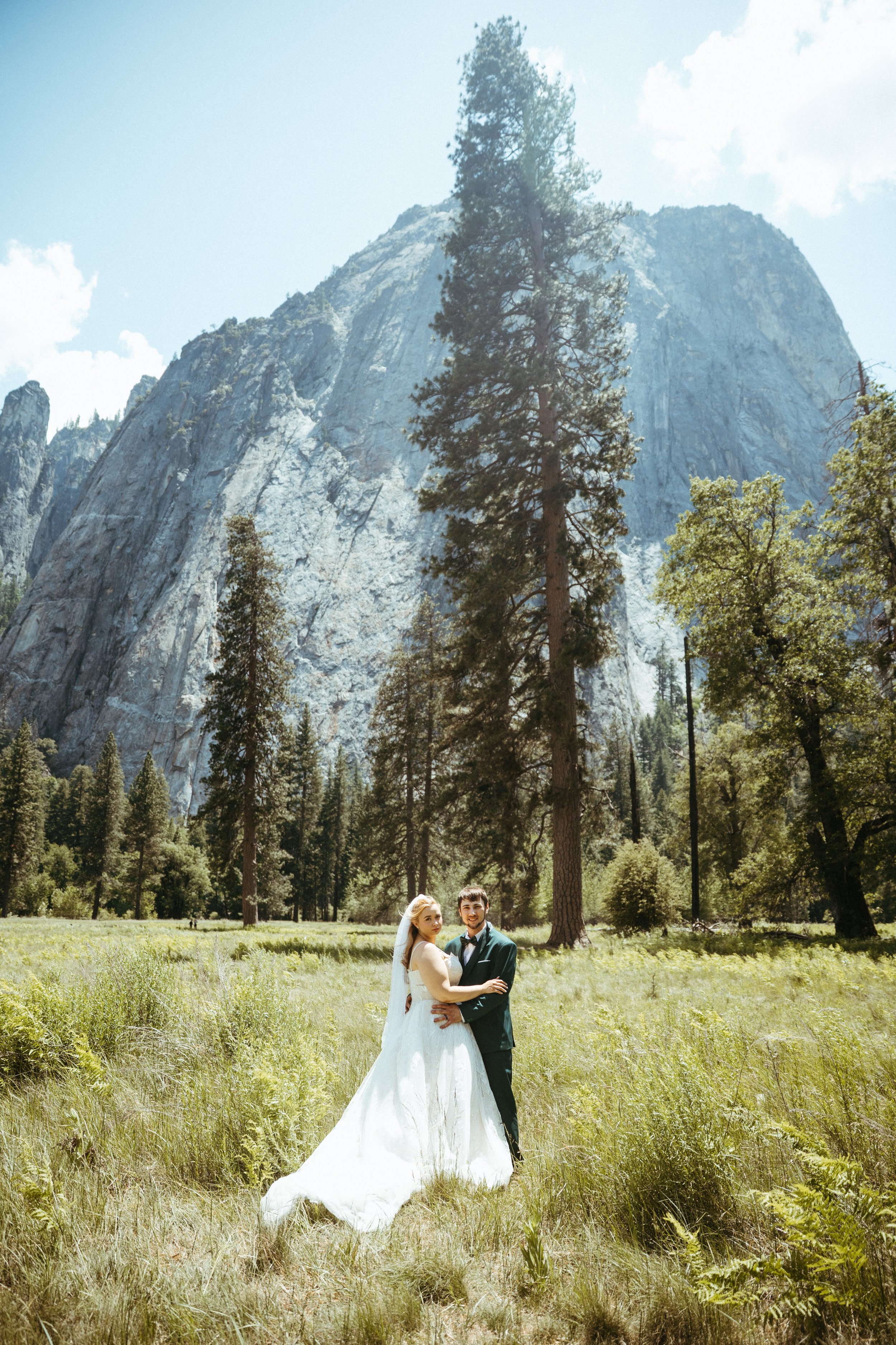 A bride and groom in wedding attire standing in a grassy clearing surrounded by tall trees, with a mountain and clear sky in the background.