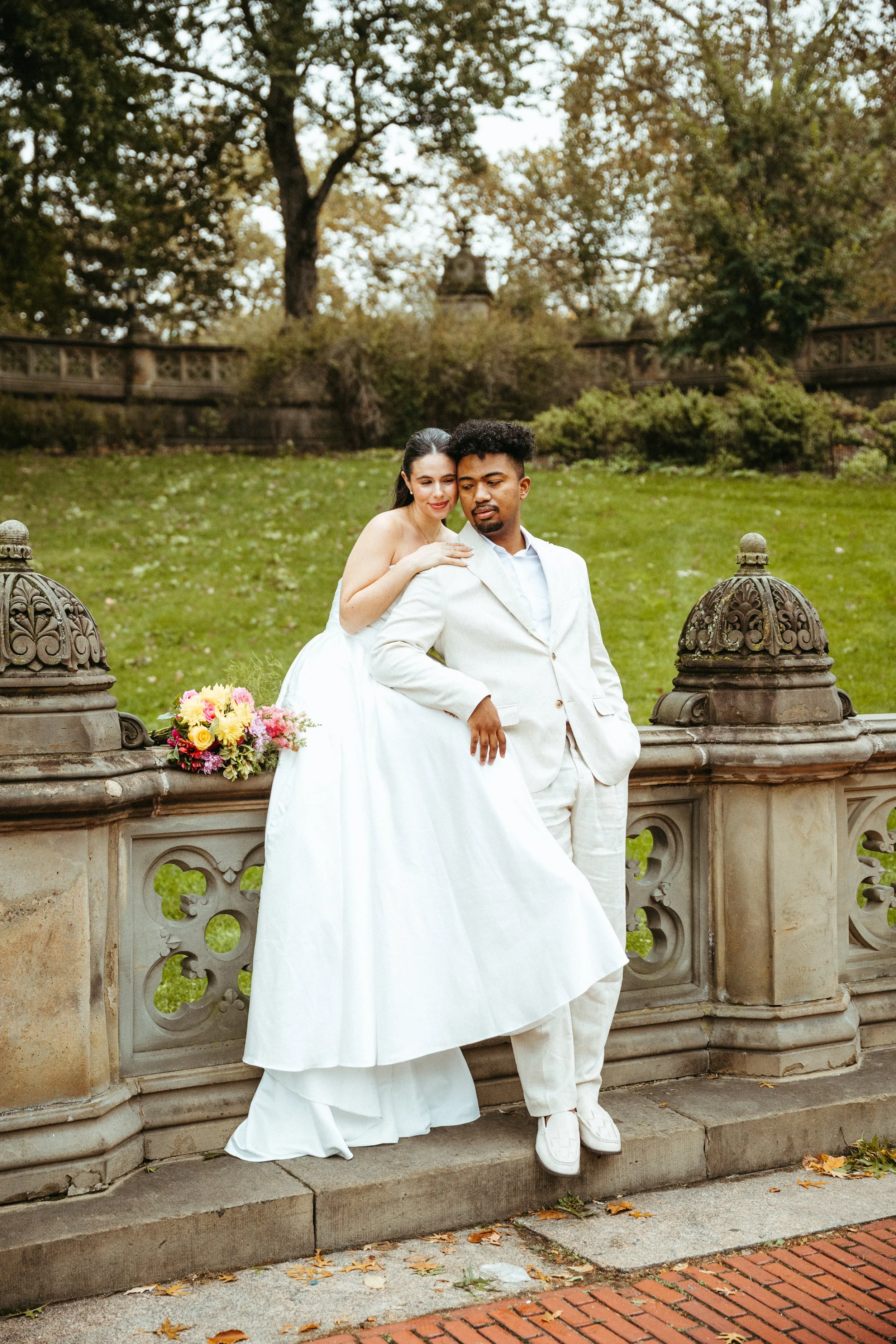 A bride and groom stand together outdoors on a stone bridge with decorative railings, surrounded by fall trees, with the bride leaning on the groom's shoulder, wearing a white wedding dress, and the groom in a light-colored suit, with a bouquet of fl