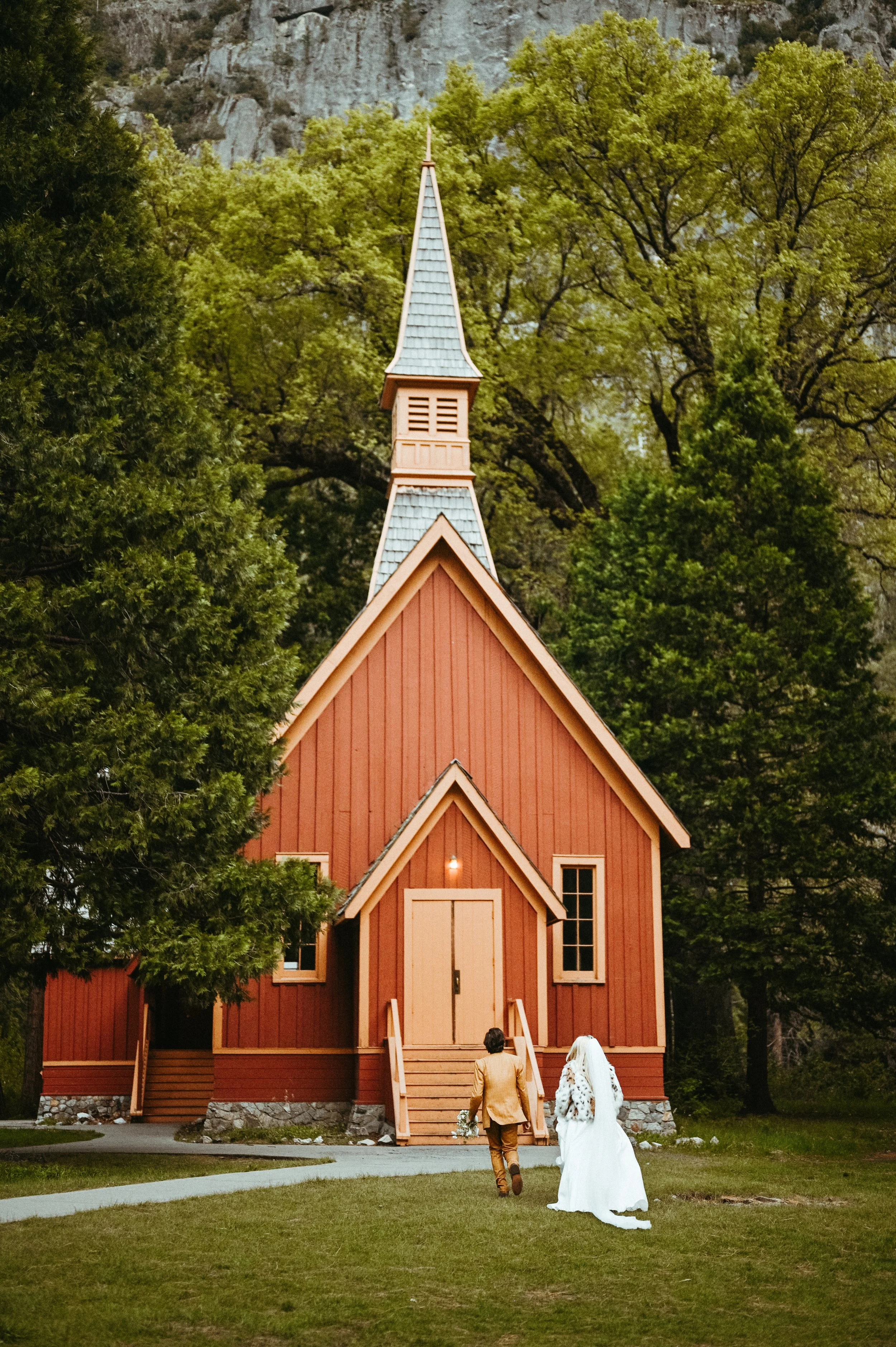 A bride in a white wedding dress and veil, and a groom in a beige suit, walking towards a small red church with a steeple, surrounded by green trees.