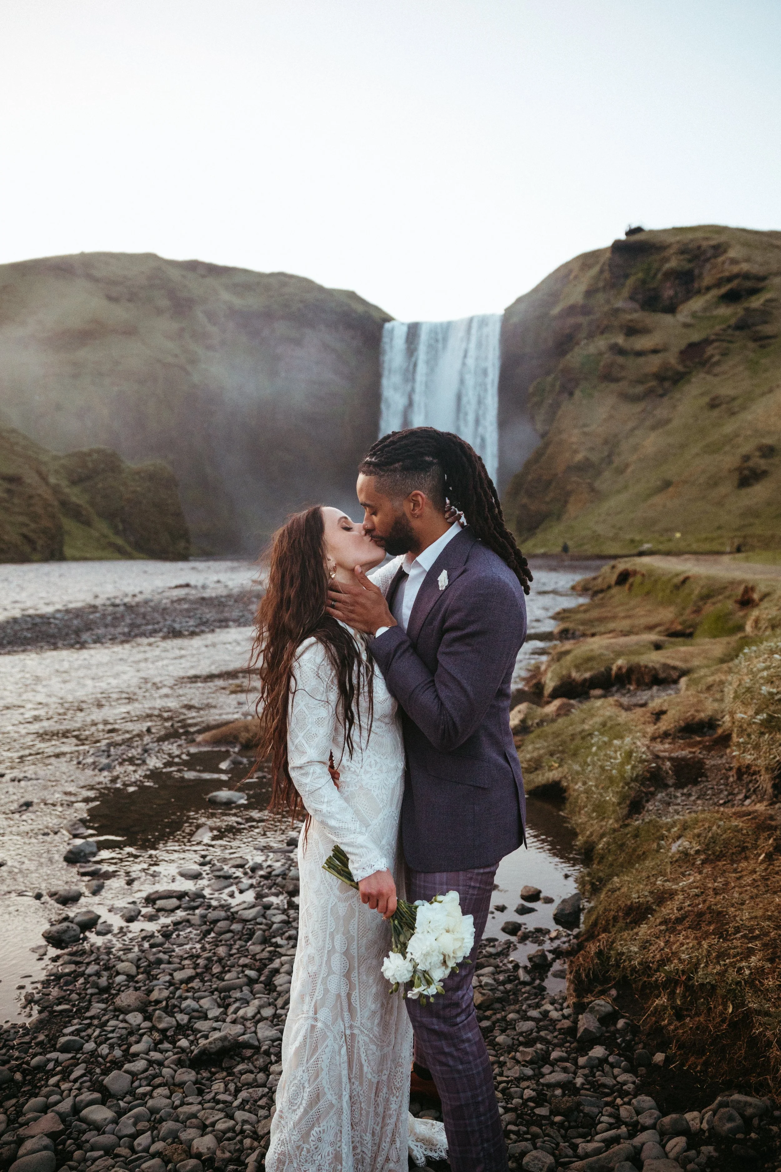 A couple in wedding attire sharing a kiss outdoors near a waterfall, with the woman holding a bouquet of white flowers.