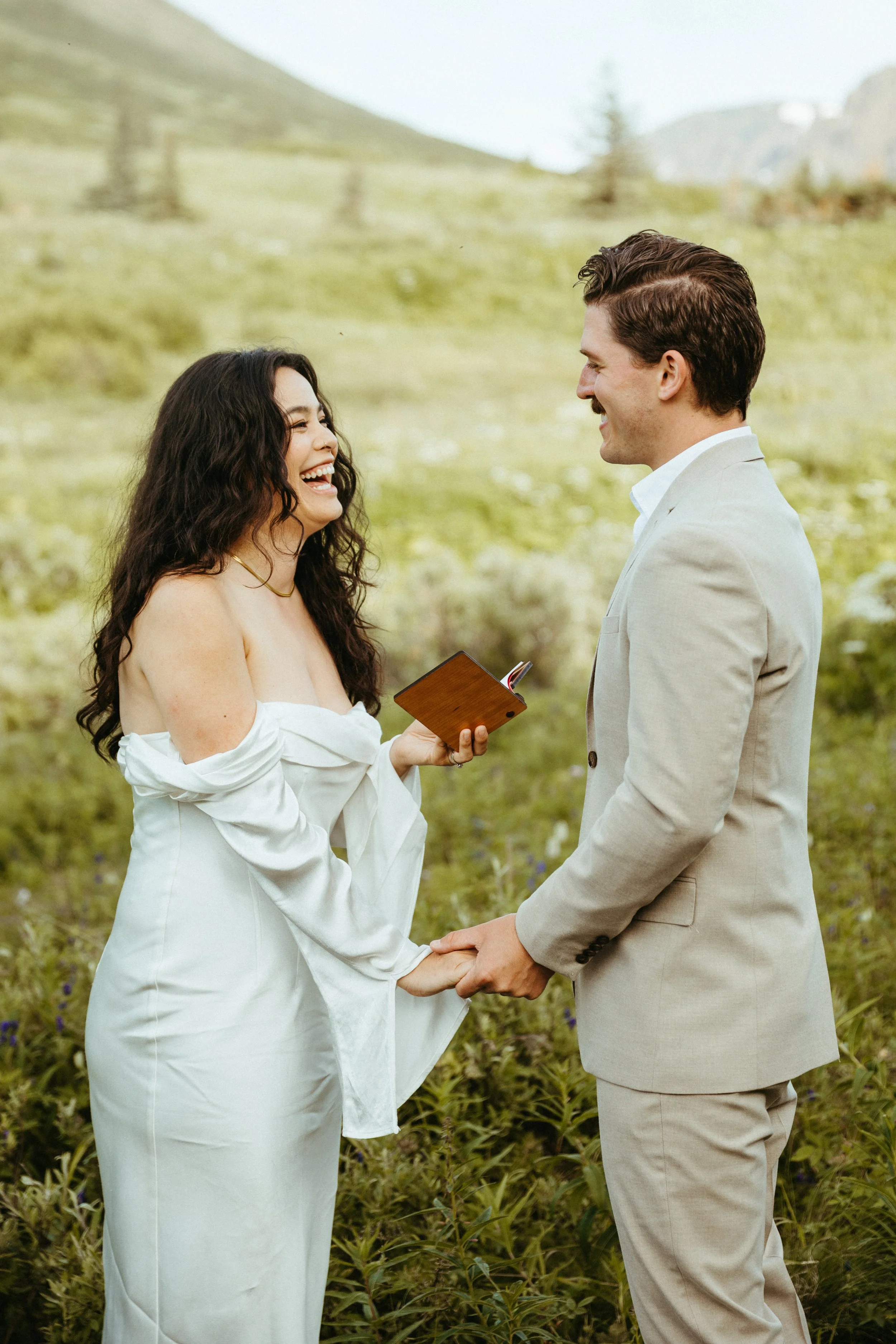 A couple holding hands and smiling during a wedding ceremony outdoors in a green hilly landscape, with the woman holding a small book.