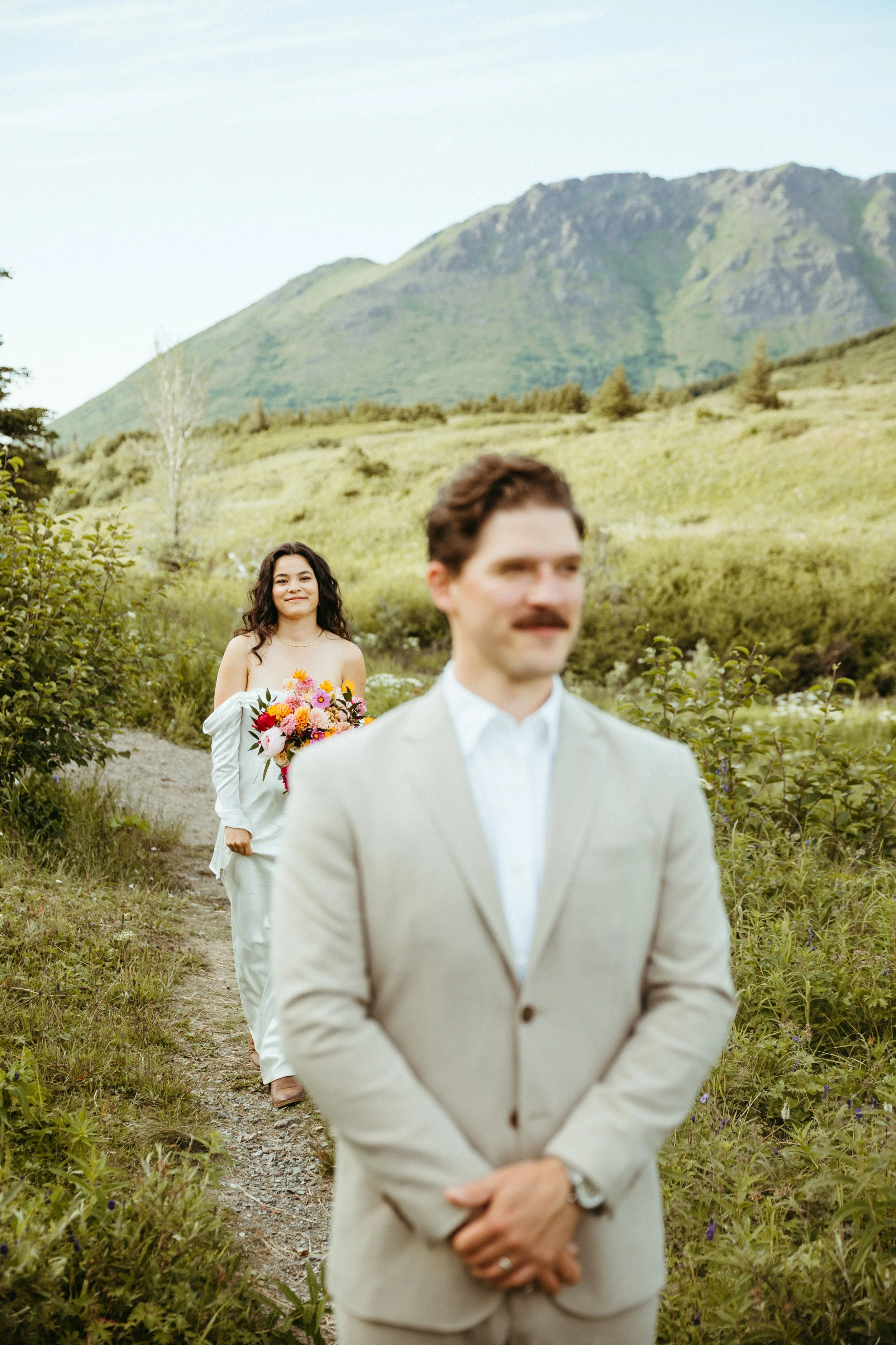 A man in a beige suit standing in the foreground with a woman in a white dress holding a colorful bouquet of flowers walking towards him in the background, in a lush green outdoor landscape with mountains in the distance.