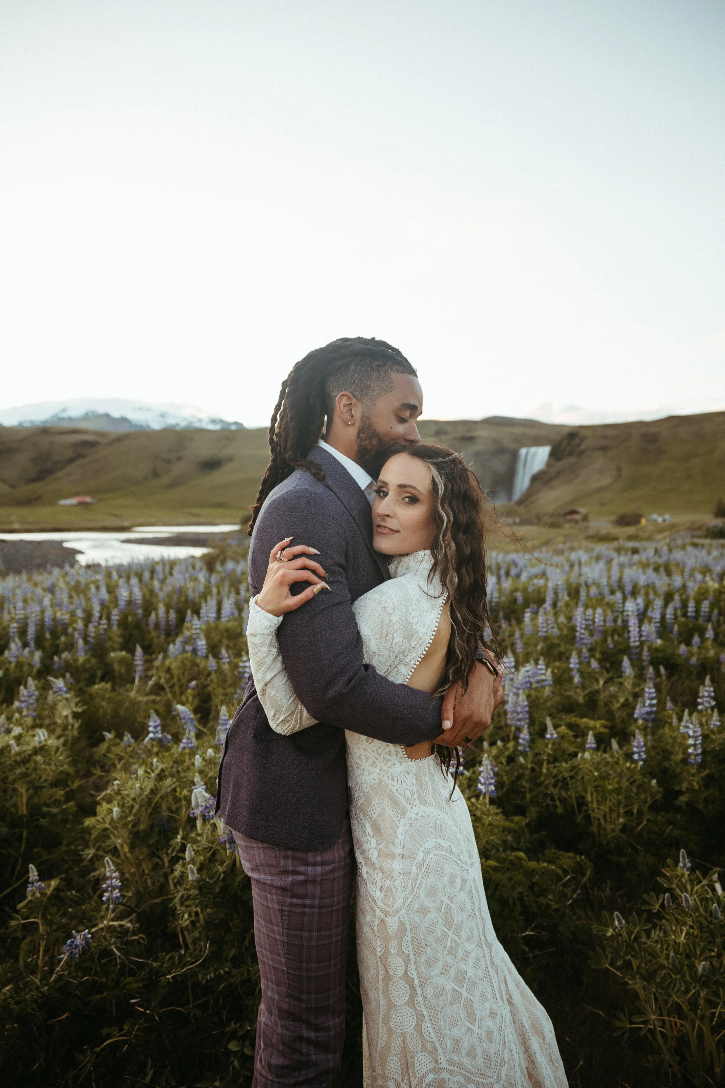 A couple embracing in a field of purple flowers with a waterfall and mountains in the background.