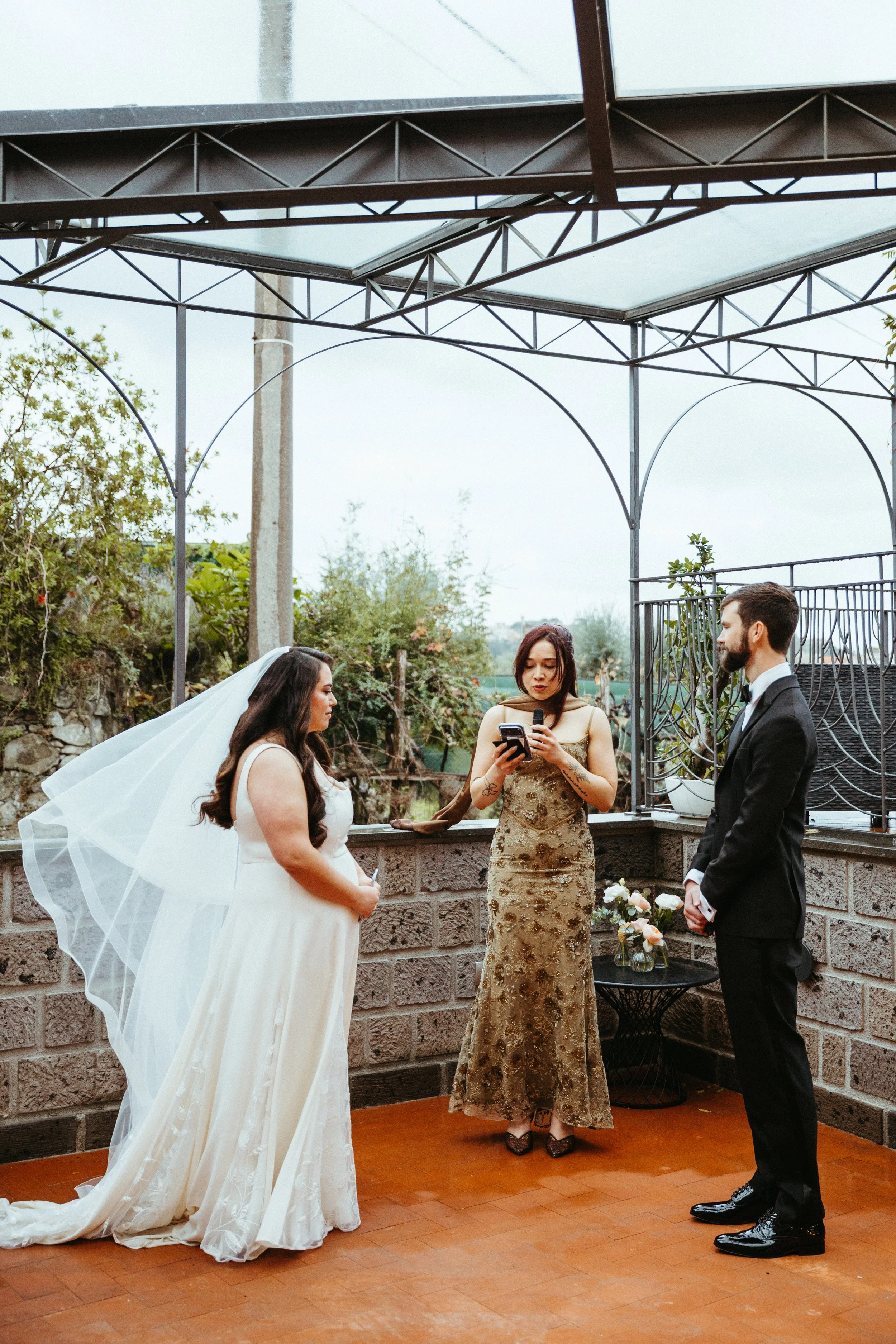 A wedding ceremony with a bride and groom standing facing each other while a woman officiates, holding a microphone and reading from a phone. The bride wears a white wedding dress and veil, and the groom wears a black tuxedo. There is a small table w