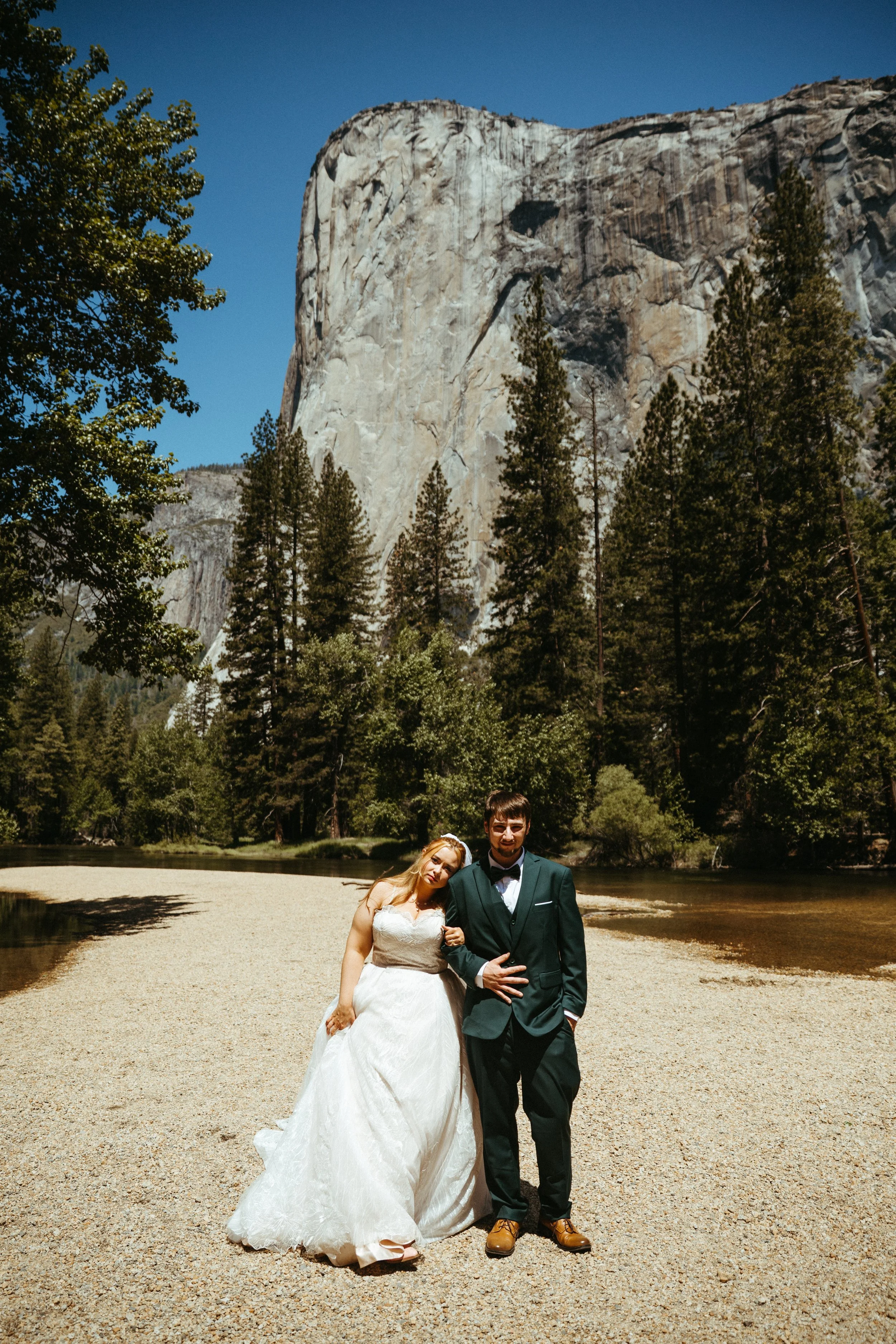 A bride in a wedding dress and a groom in a tuxedo standing arm-in-arm on a gravel area near a river with mountains and trees in the background.