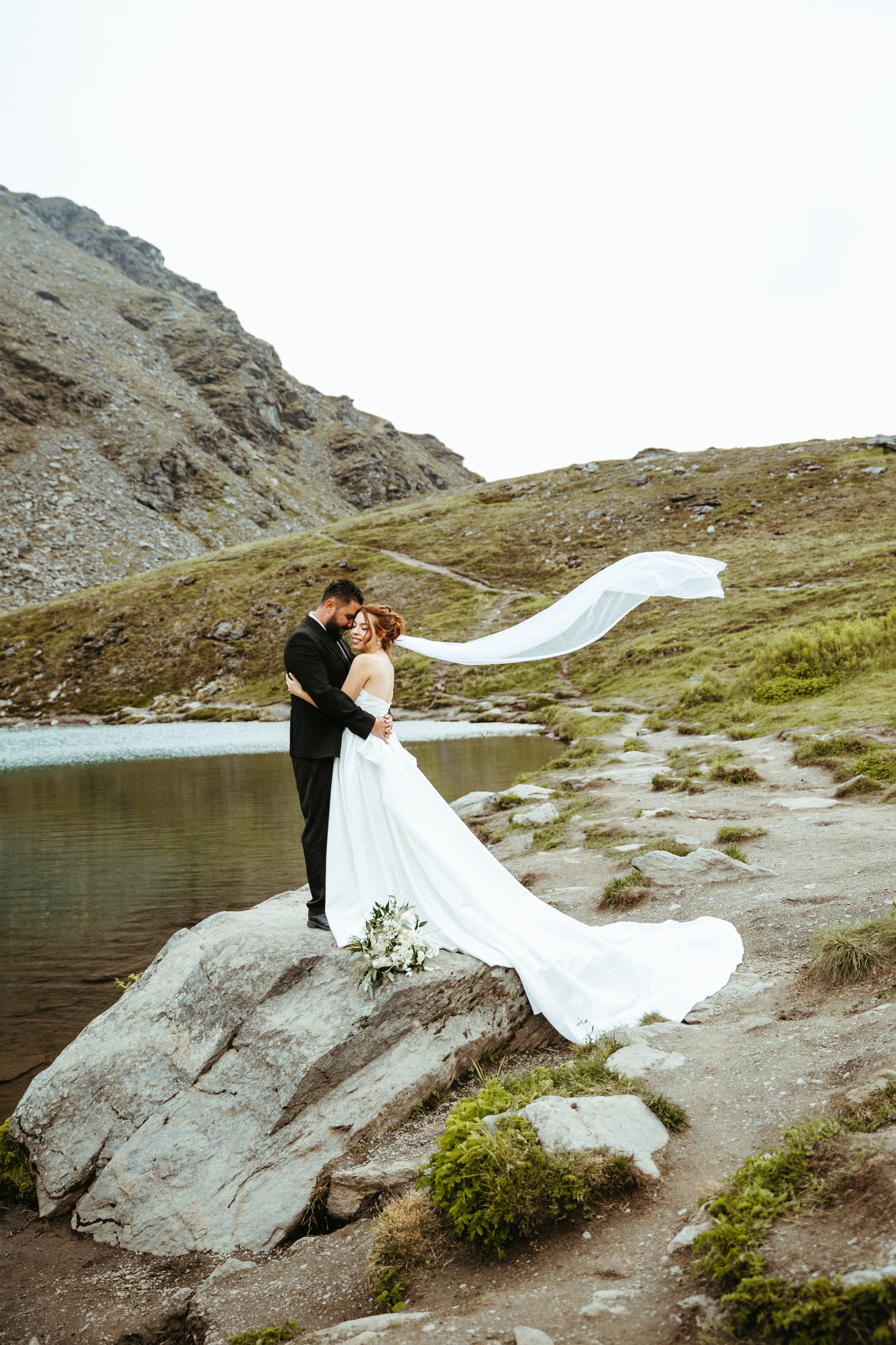 A bride and groom standing on a large rock near a lake with mountainous terrain in the background, embracing each other with the bride’s veil flowing in the wind.
