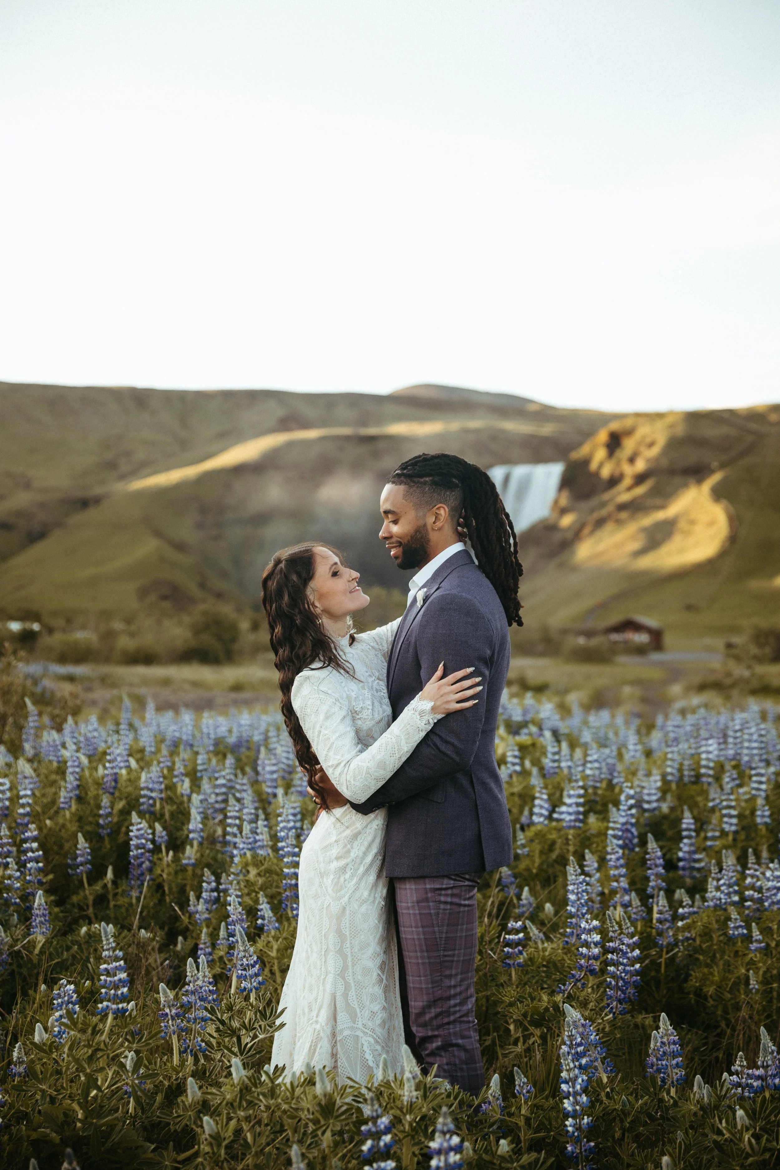 A couple embracing in a field of purple flowers with a waterfall and mountains in the background.