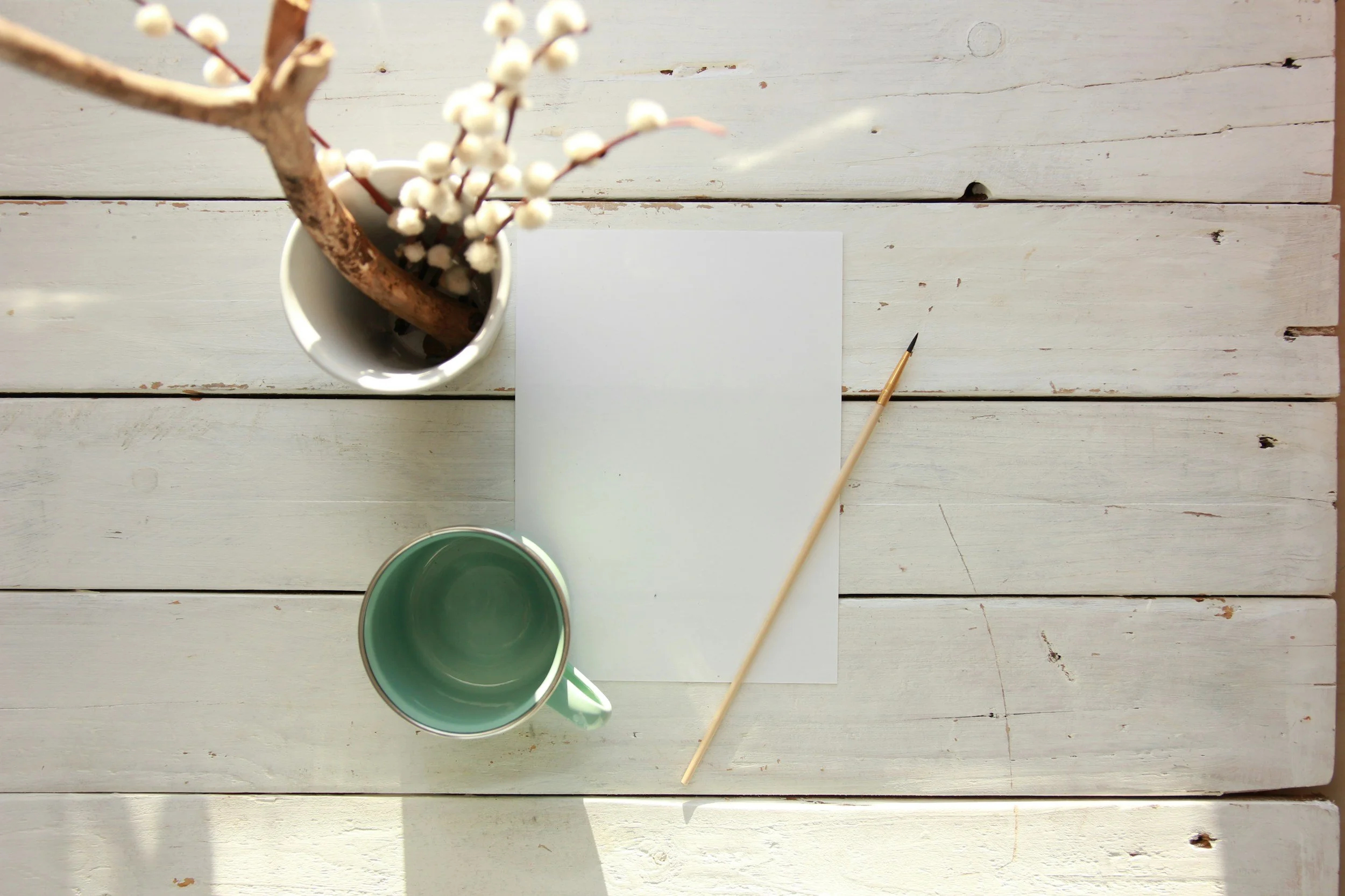 A white wooden table with a blank sheet of paper, a paintbrush, a green cup, and a vase with a branch of white flowers.