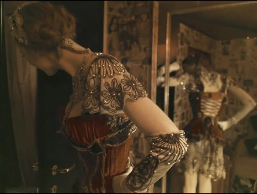 A woman in ornate, vintage-style clothing inside a room with patterned wallpaper, looking at herself in a mirror.